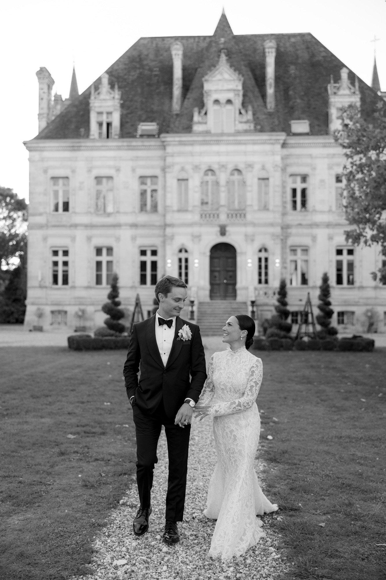 A bride and groom in formal attire walk hand in hand on a gravel path in front of a large, ornate building.