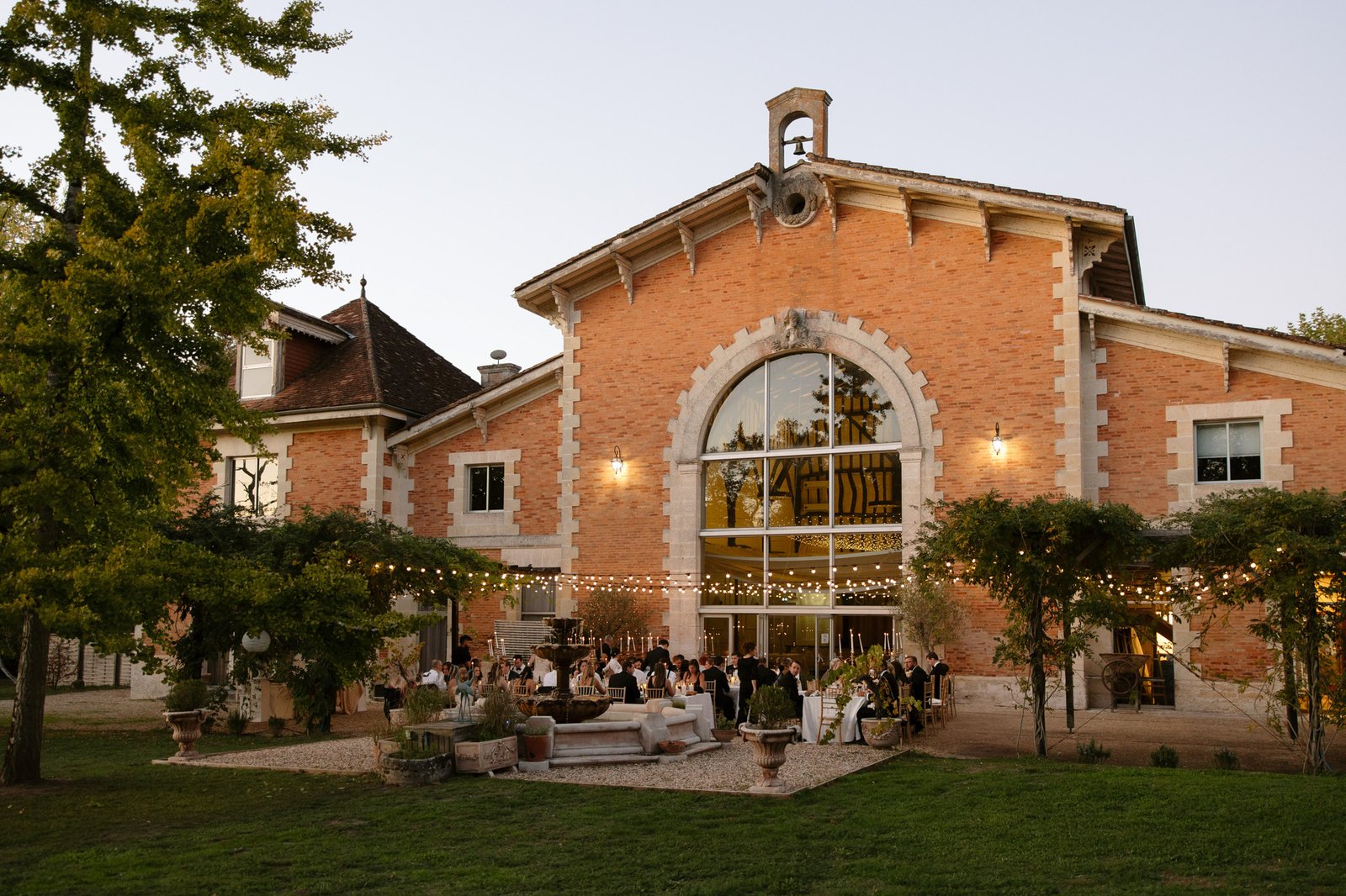 A group of people dine outdoors under string lights beside a large brick building with arched windows and greenery at dusk.