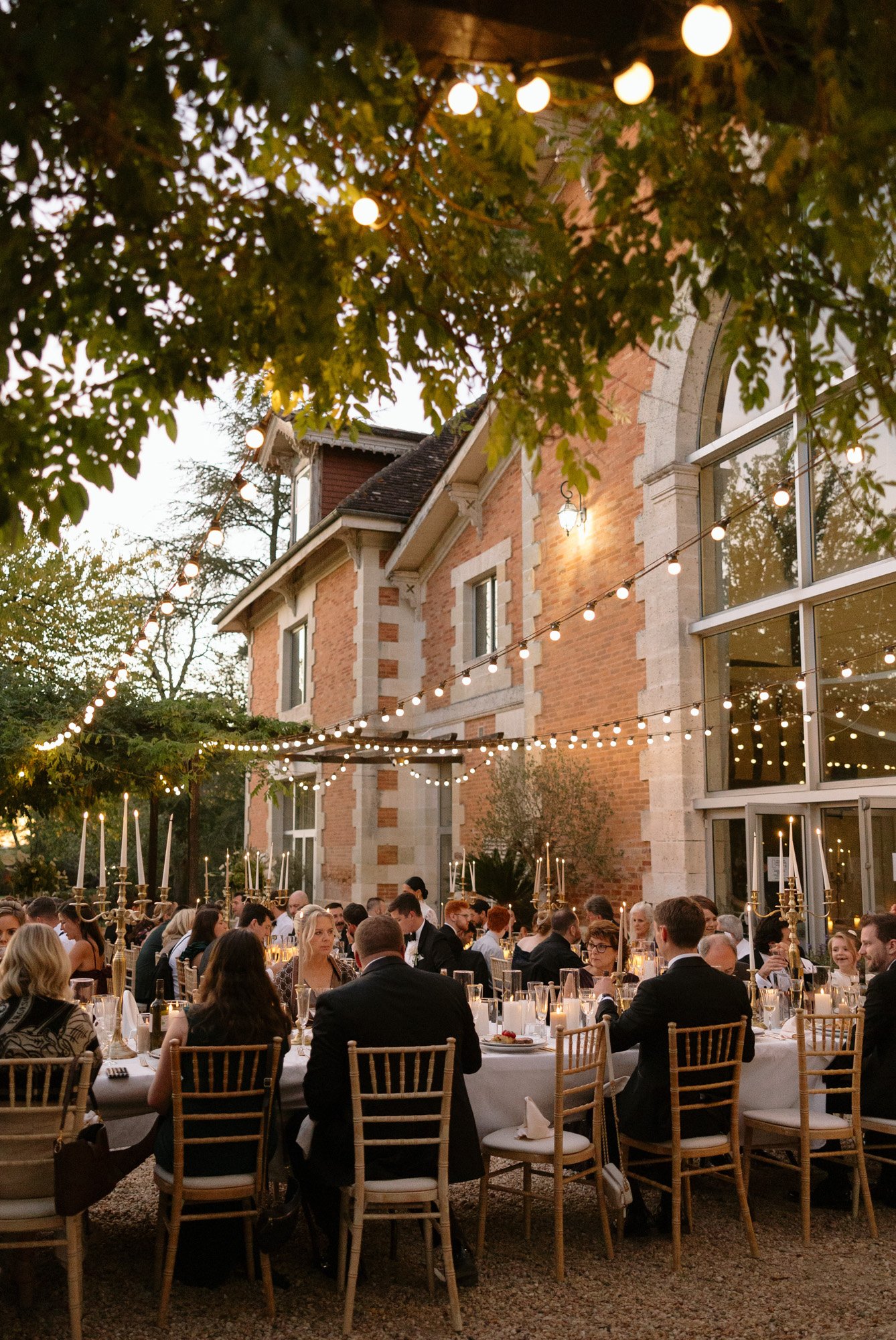 People seated around a long outdoor dining table under string lights in front of a brick building, with trees and candle decorations visible. Chateau de la Valouze wedding.