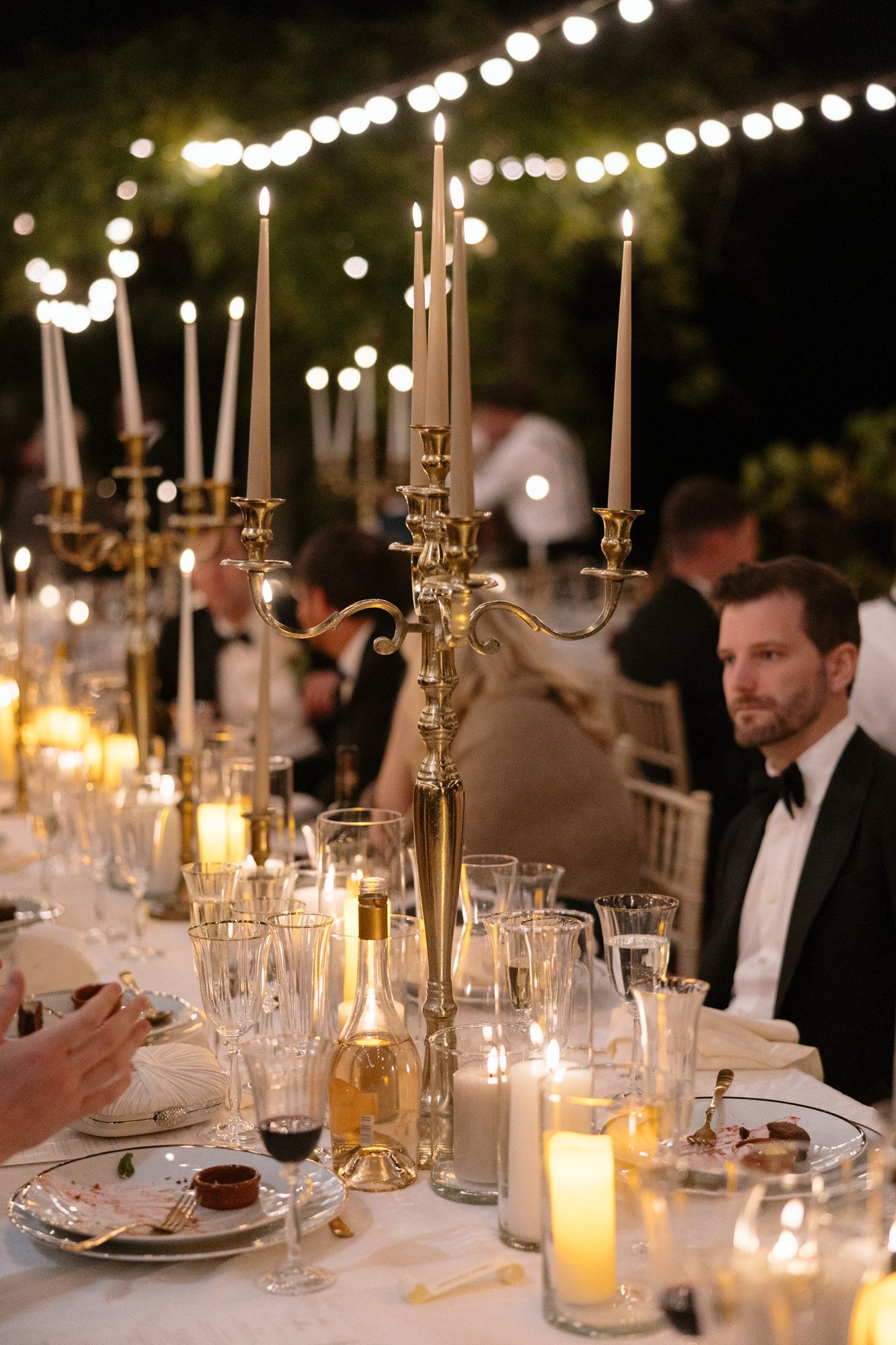A formal outdoor dinner table is set with candles, glassware, and candelabras; guests in formal attire are seated, and string lights hang above.