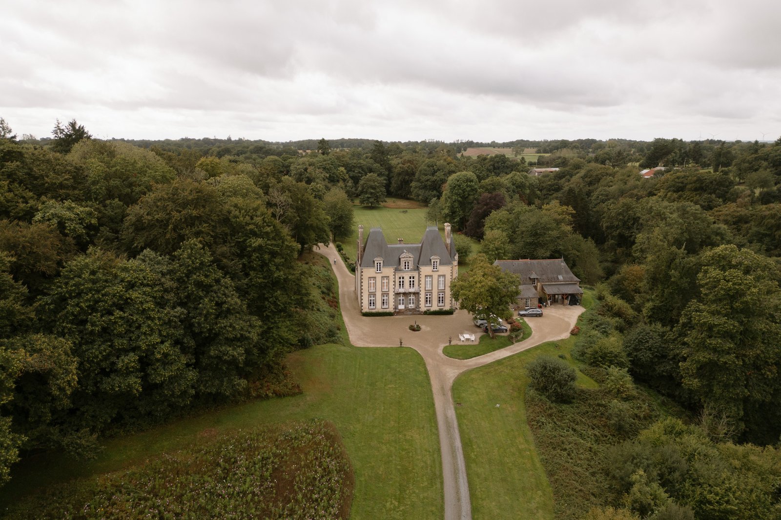 A large, old-style house with a dark roof is surrounded by trees and greenery, with a driveway leading to the main entrance and another building nearby.