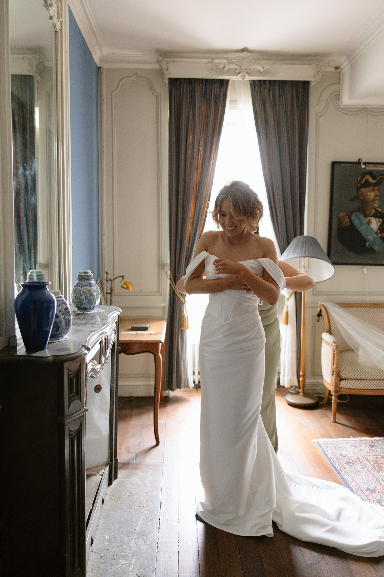 A woman in a white wedding dress is being assisted by another person in a well-lit, elegant room with classic decor and a portrait on the wall.