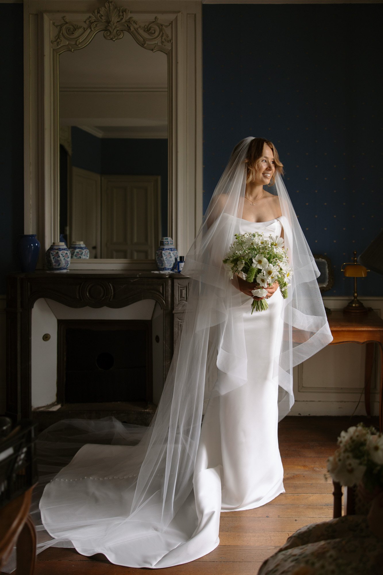 A bride in a white wedding dress and long veil stands indoors holding a bouquet of flowers, with a fireplace and mirror in the background.