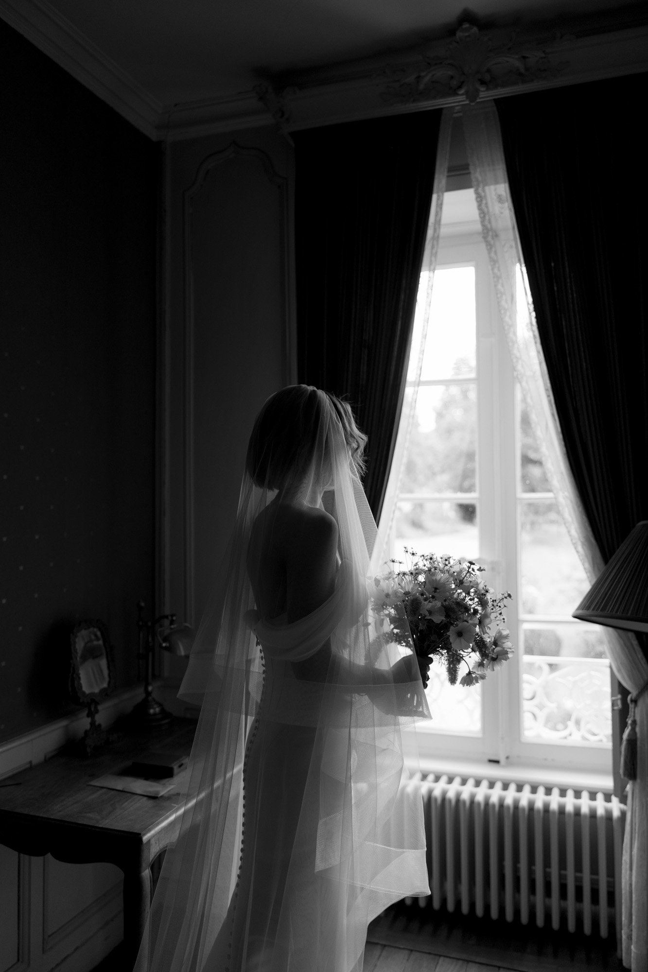A bride in a wedding dress and veil stands by a window holding a bouquet, with light streaming in through the curtains.