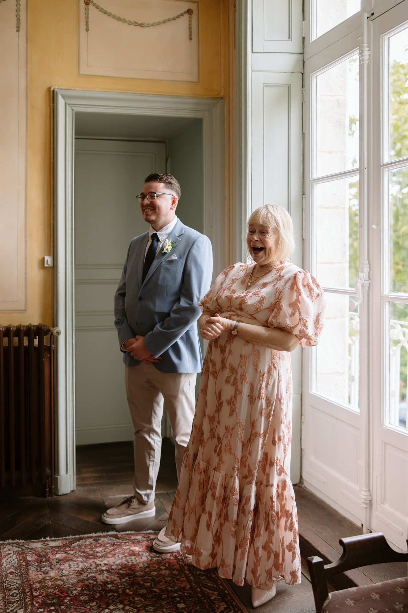 A man in a blue suit and a woman in a patterned dress stand indoors by tall windows, both smiling and appearing joyful.