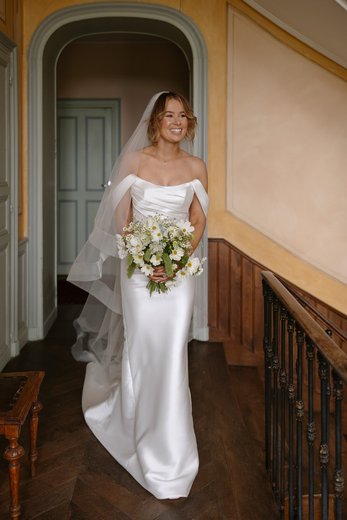A bride in a white off-shoulder gown and veil stands indoors, holding a bouquet of white and yellow flowers, smiling.