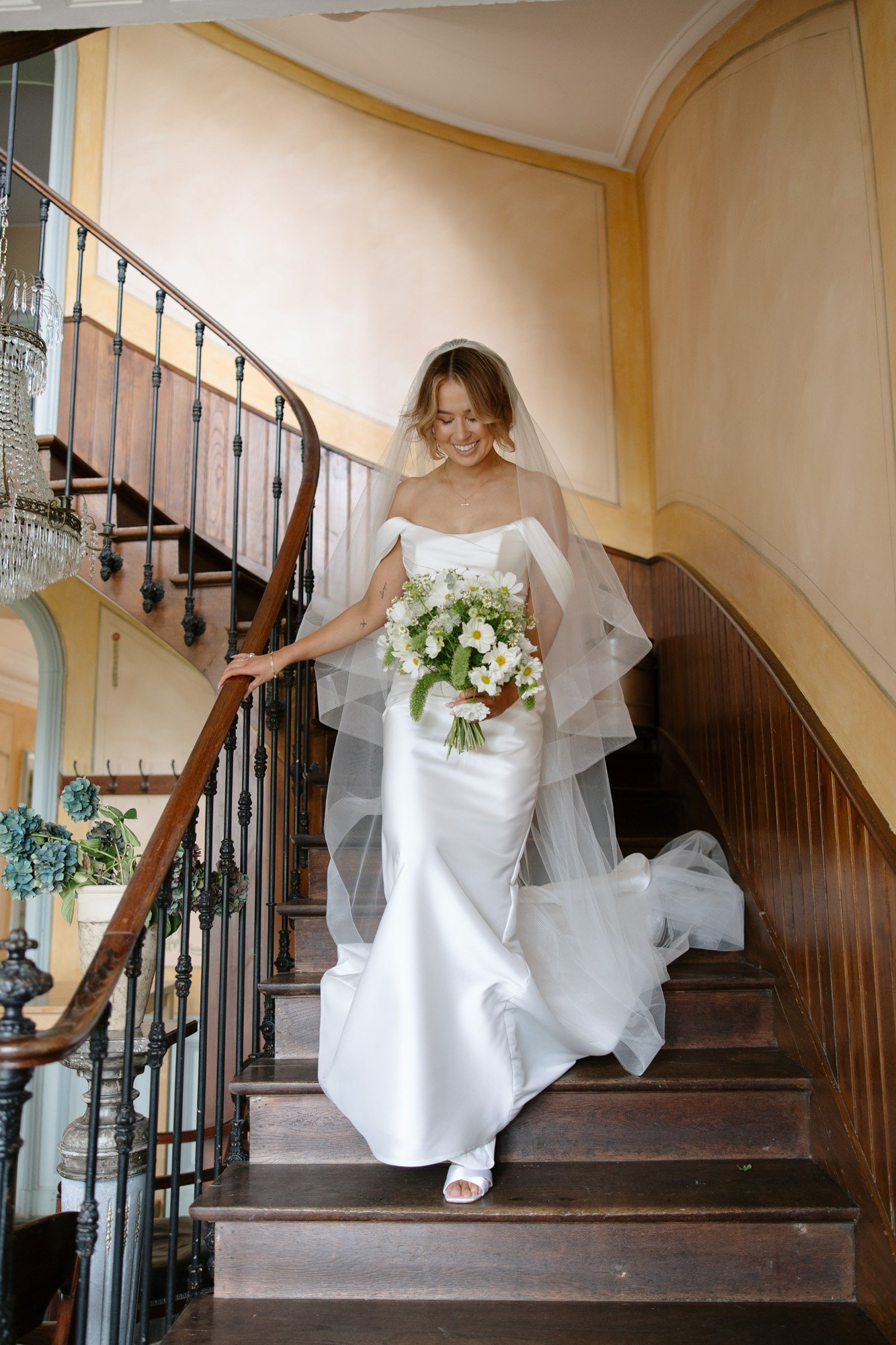 A bride in a white gown and veil descends a wooden staircase, holding a bouquet of flowers.