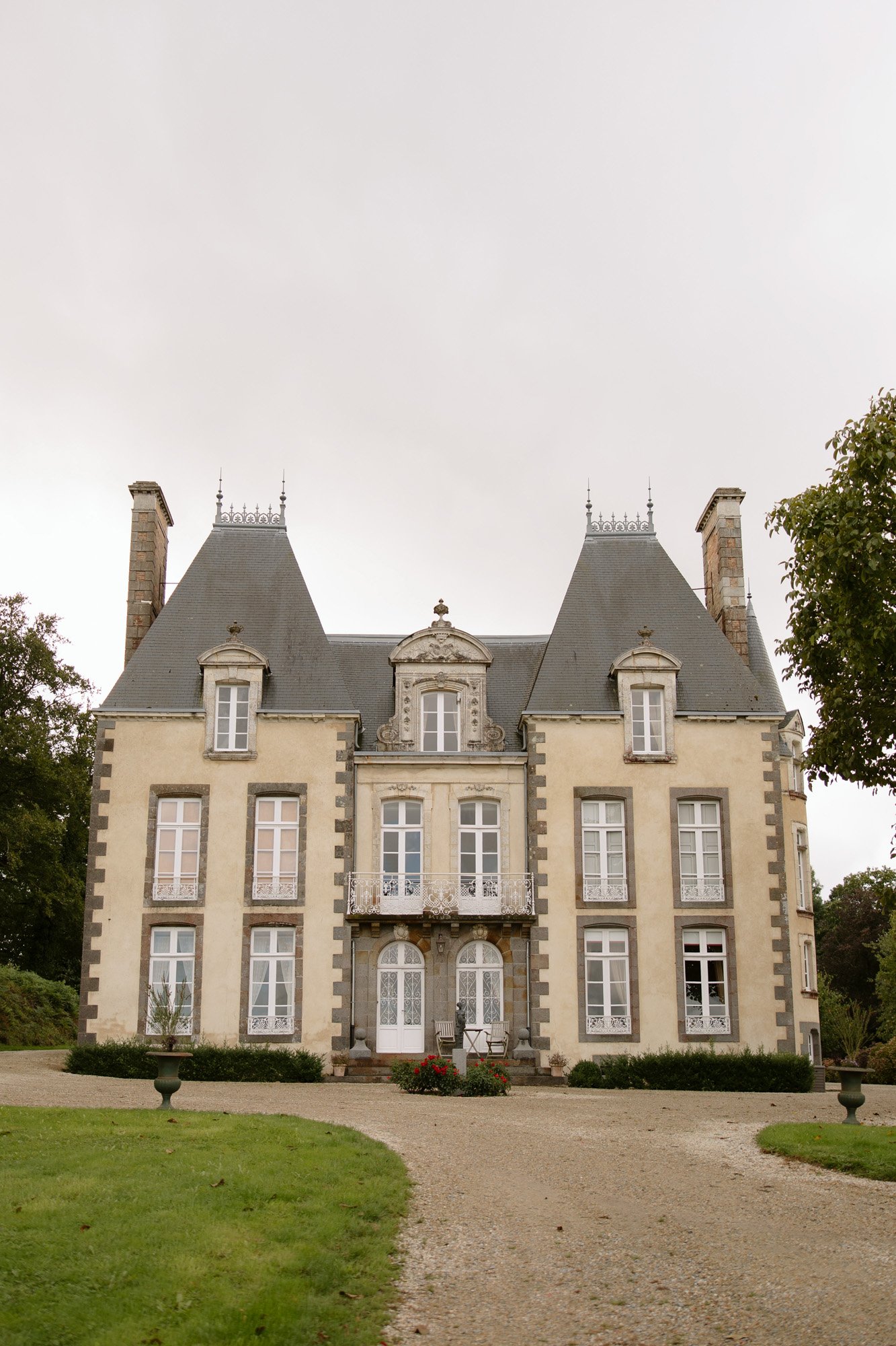 A beige, three-story French chateau with steep slate roofs, tall windows, and ornate ironwork, set against an overcast sky with trees and grass in the foreground.