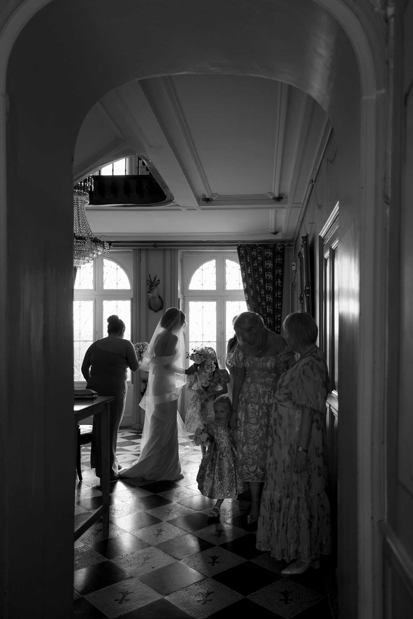 A bride and several women, some holding a child, stand together in a hallway with large windows and patterned floor tiles.