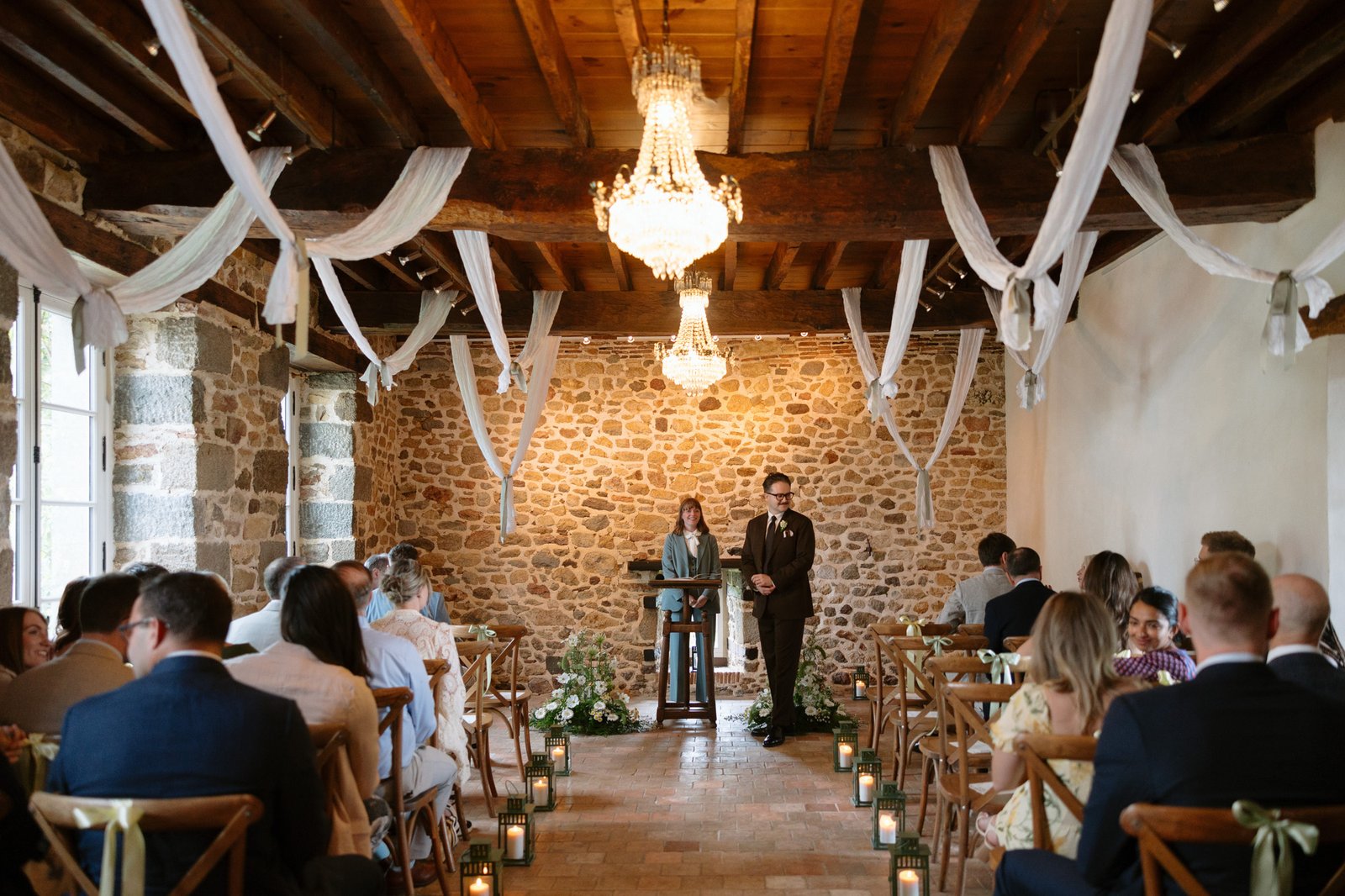 Indoor wedding ceremony with guests seated, a stone wall backdrop, chandeliers above, and an officiant and man standing at the front near flower arrangements.