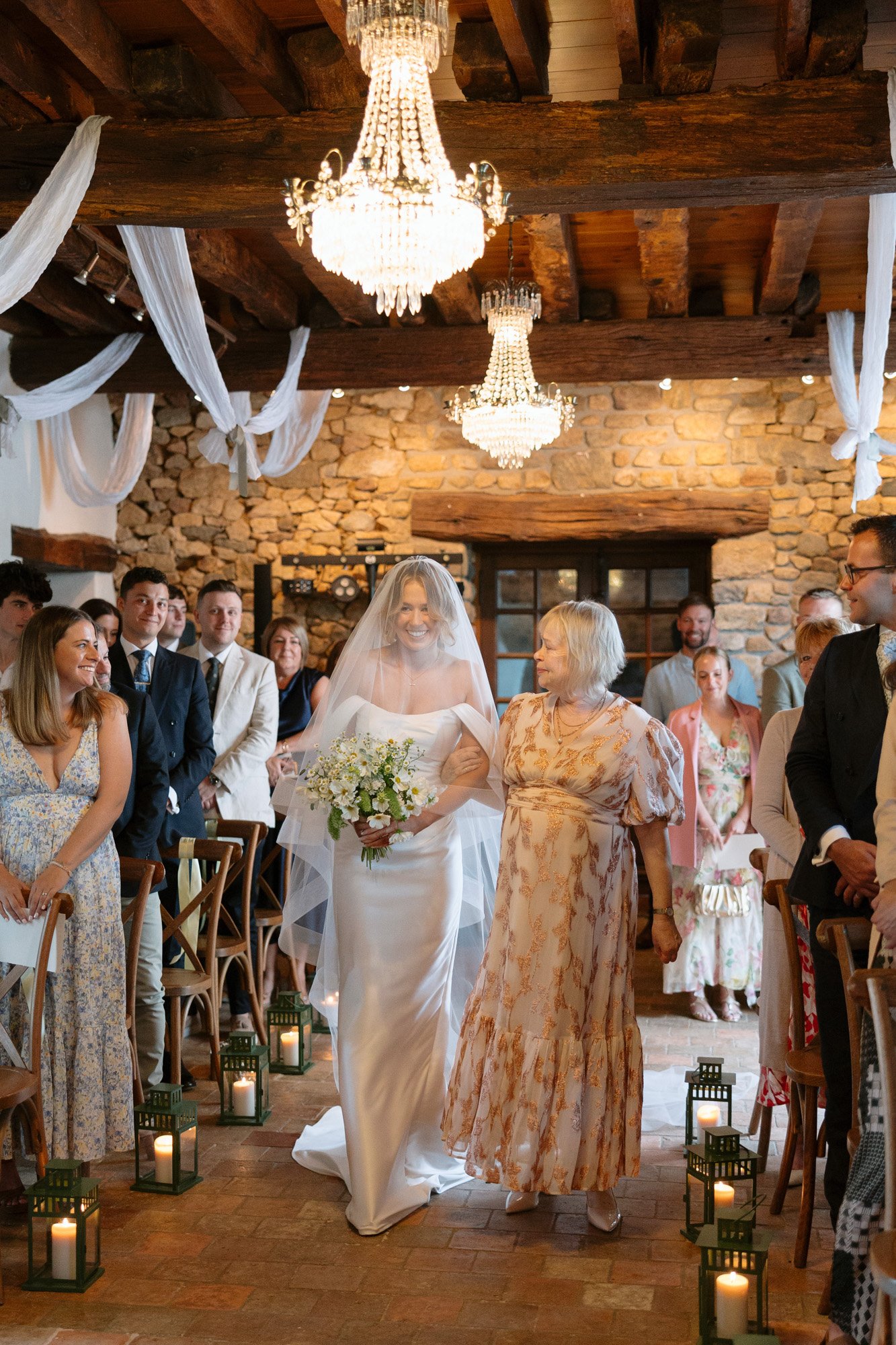 A bride in a white dress and veil walks down the aisle with an older woman beside her, surrounded by seated and standing guests in a rustic venue decorated with chandeliers and lanterns.