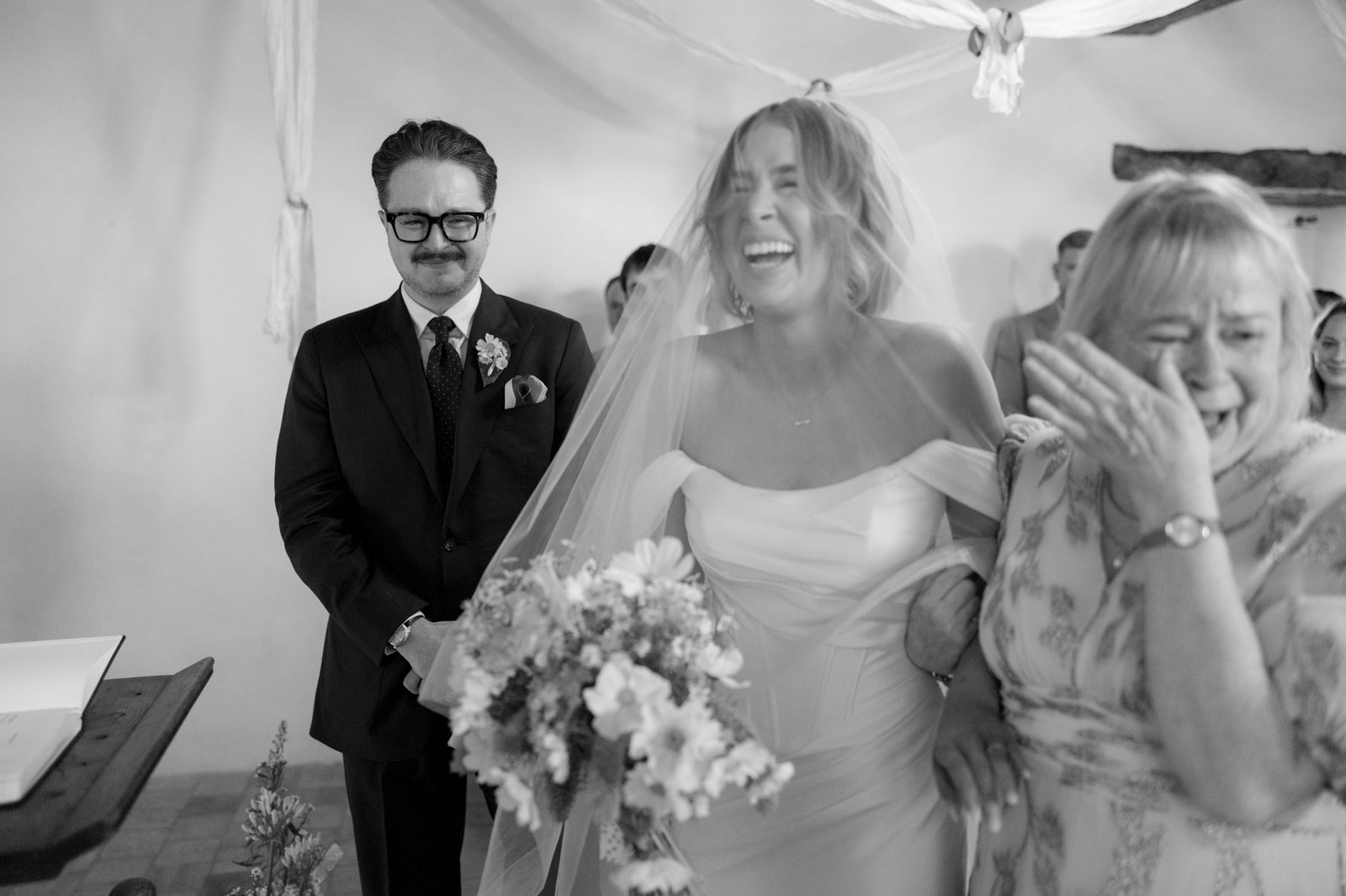A bride in a wedding dress laughs joyfully, holding a bouquet and arm-in-arm with an emotional woman, while a man in a suit stands beside them, smiling.