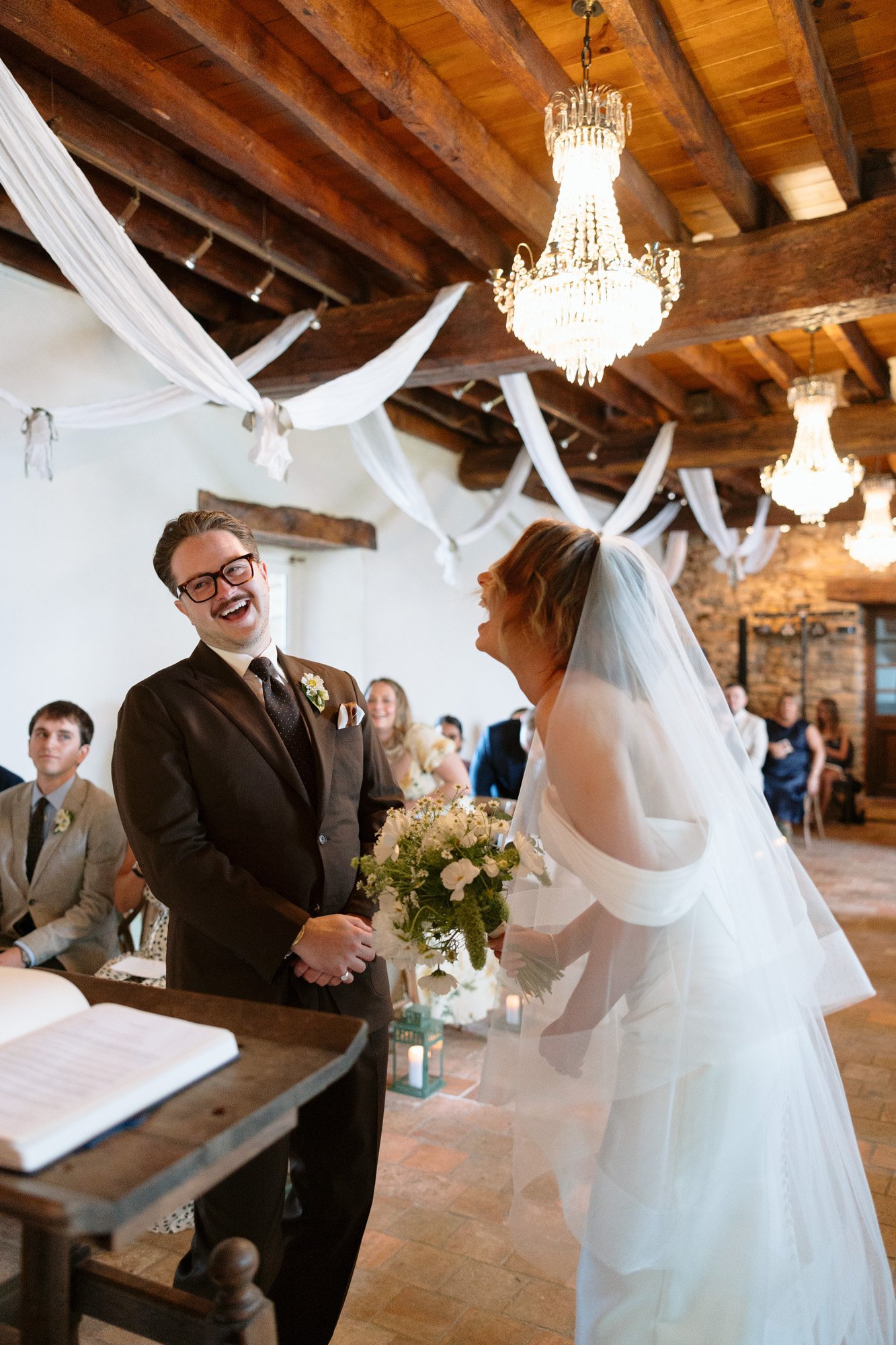 A bride and groom stand at the altar, smiling and laughing, during a wedding ceremony in a rustic, decorated hall with chandeliers and wooden beams.