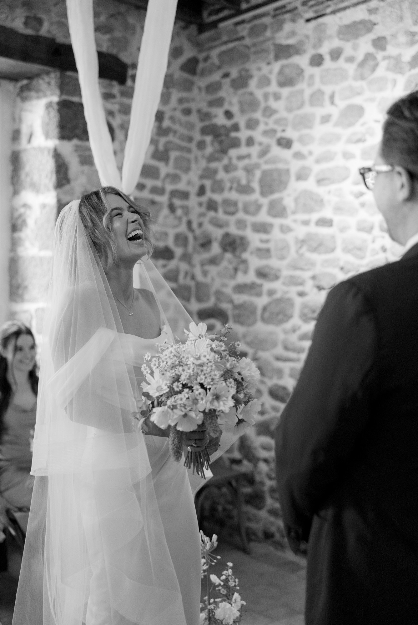 A bride in a veil and holding a bouquet smiles at a person in a suit during a wedding ceremony in a stone-walled room.