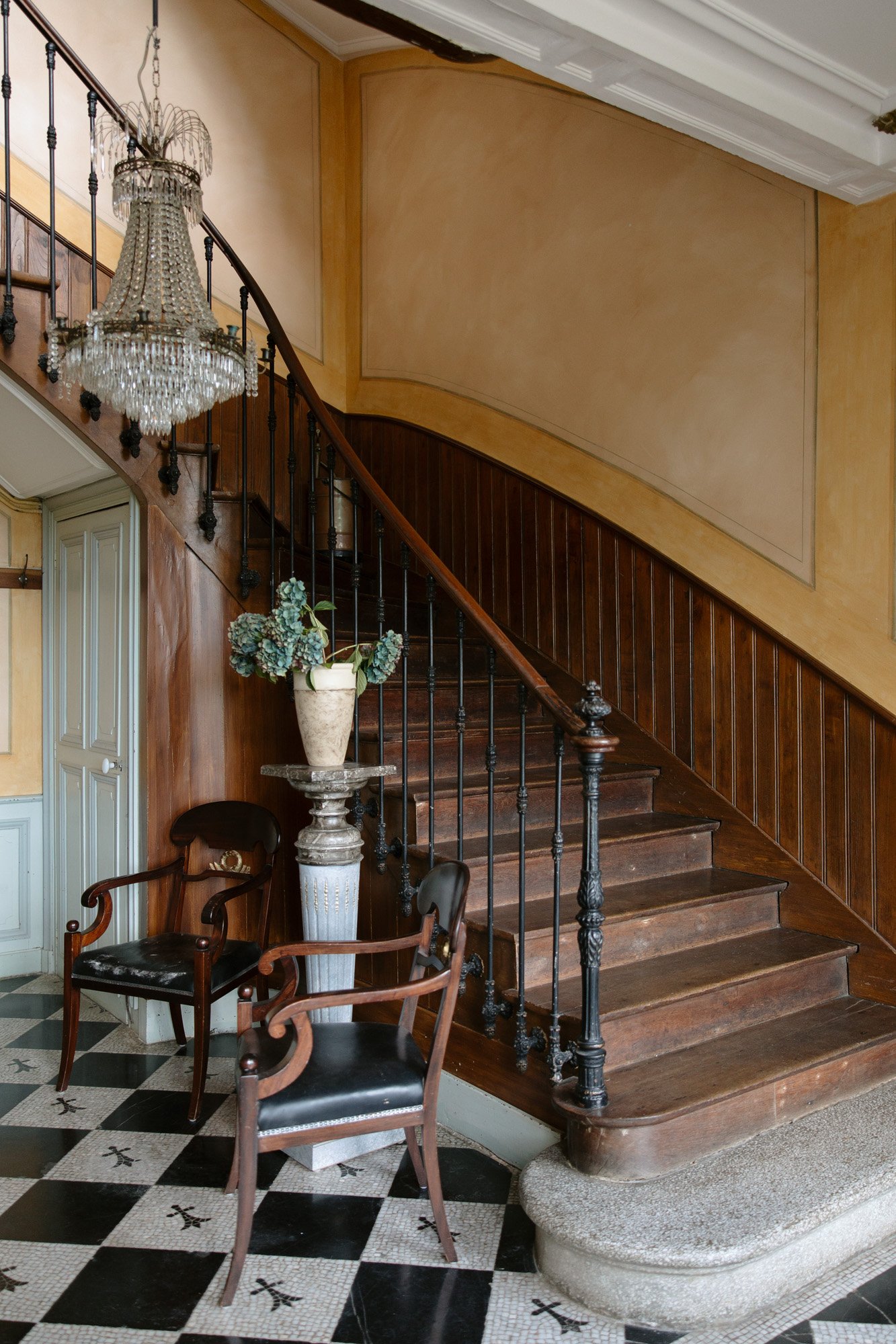 A curved wooden staircase with a black metal railing, a crystal chandelier, two wooden chairs, a vase with greenery, and a black-and-white checkered tile floor.