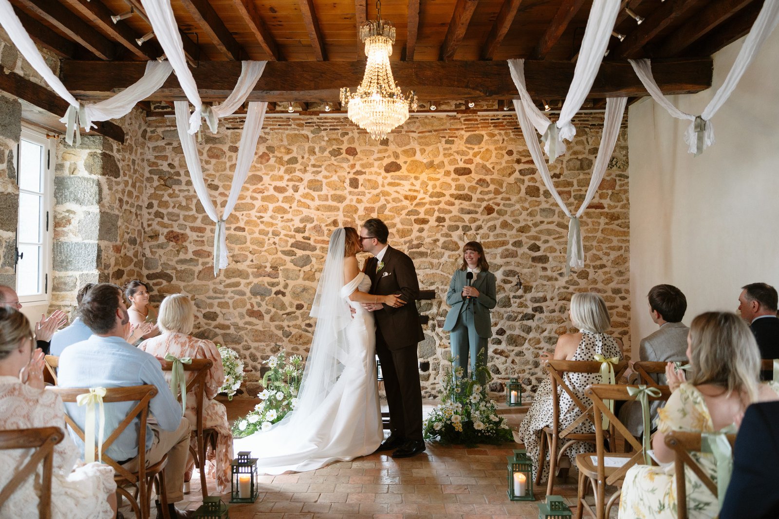 A bride and groom kiss at the altar during a wedding ceremony, surrounded by seated guests in a rustic, stone-walled venue with white drapery and floral decorations.
