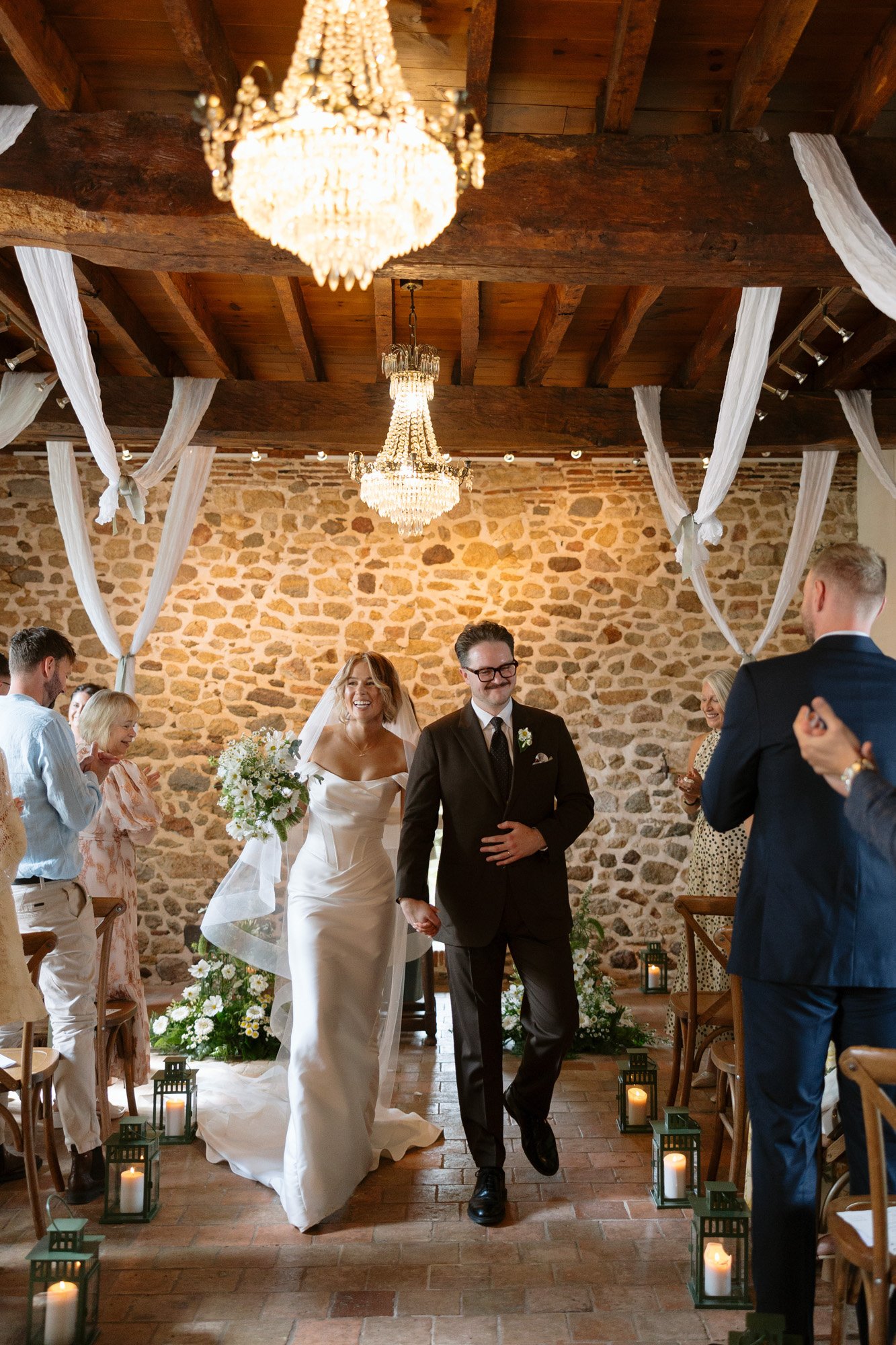A bride and groom walk down the aisle together in a rustic indoor venue with stone walls, chandeliers, and guests seated on either side, clapping.