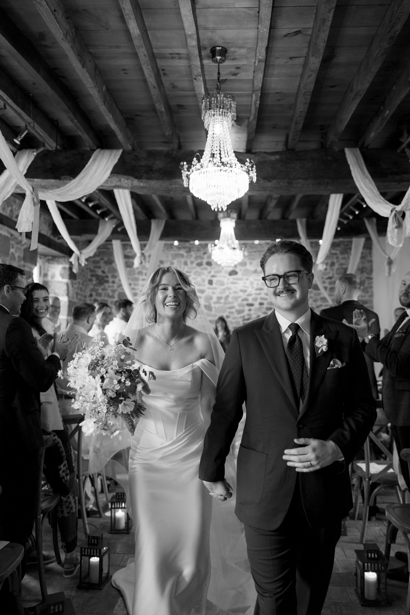 A bride and groom walk down the aisle holding hands in a decorated indoor venue, surrounded by guests and lit by a chandelier.