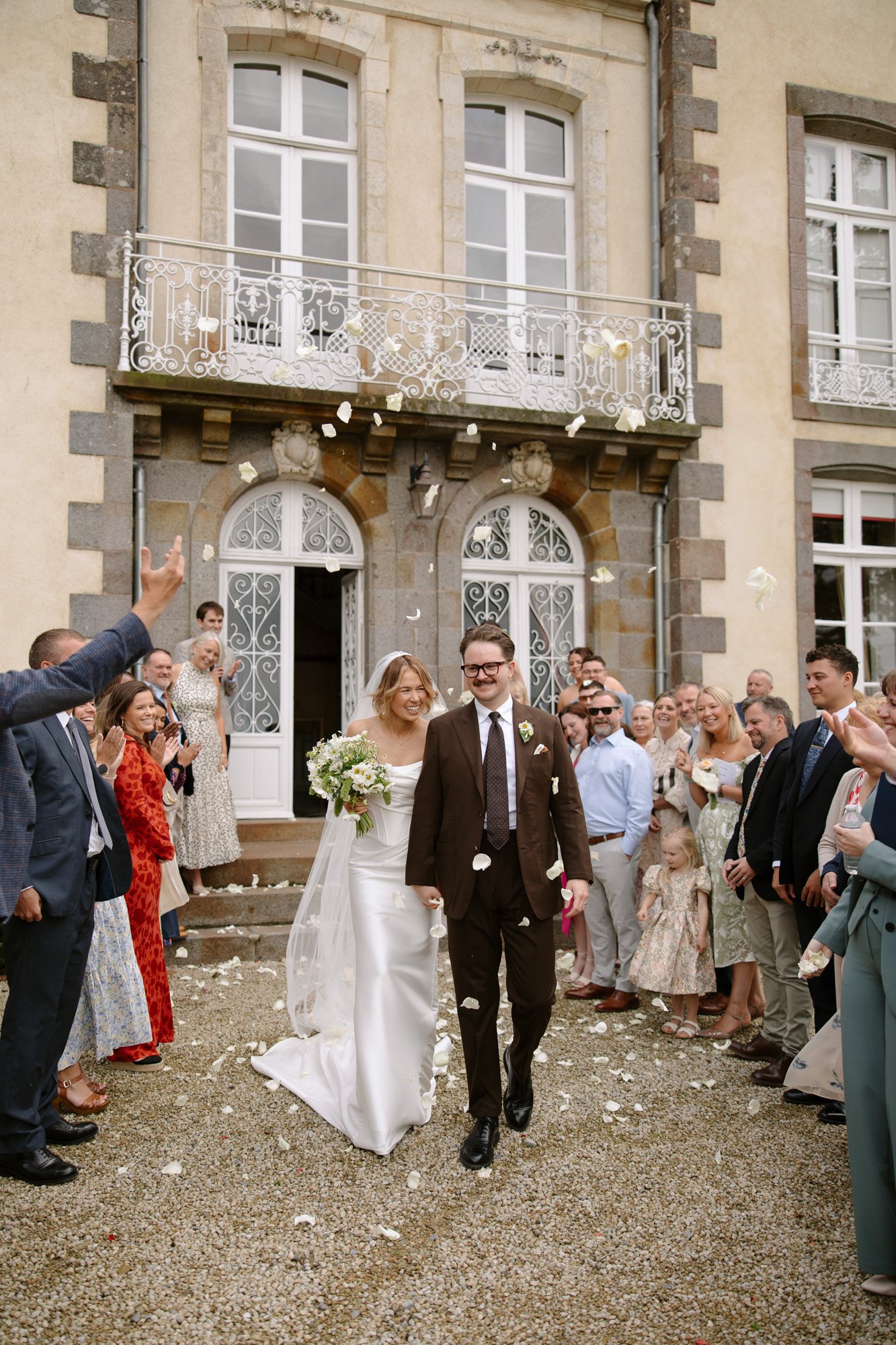 A bride and groom walk arm in arm outside a building, surrounded by guests throwing flower petals in celebration.