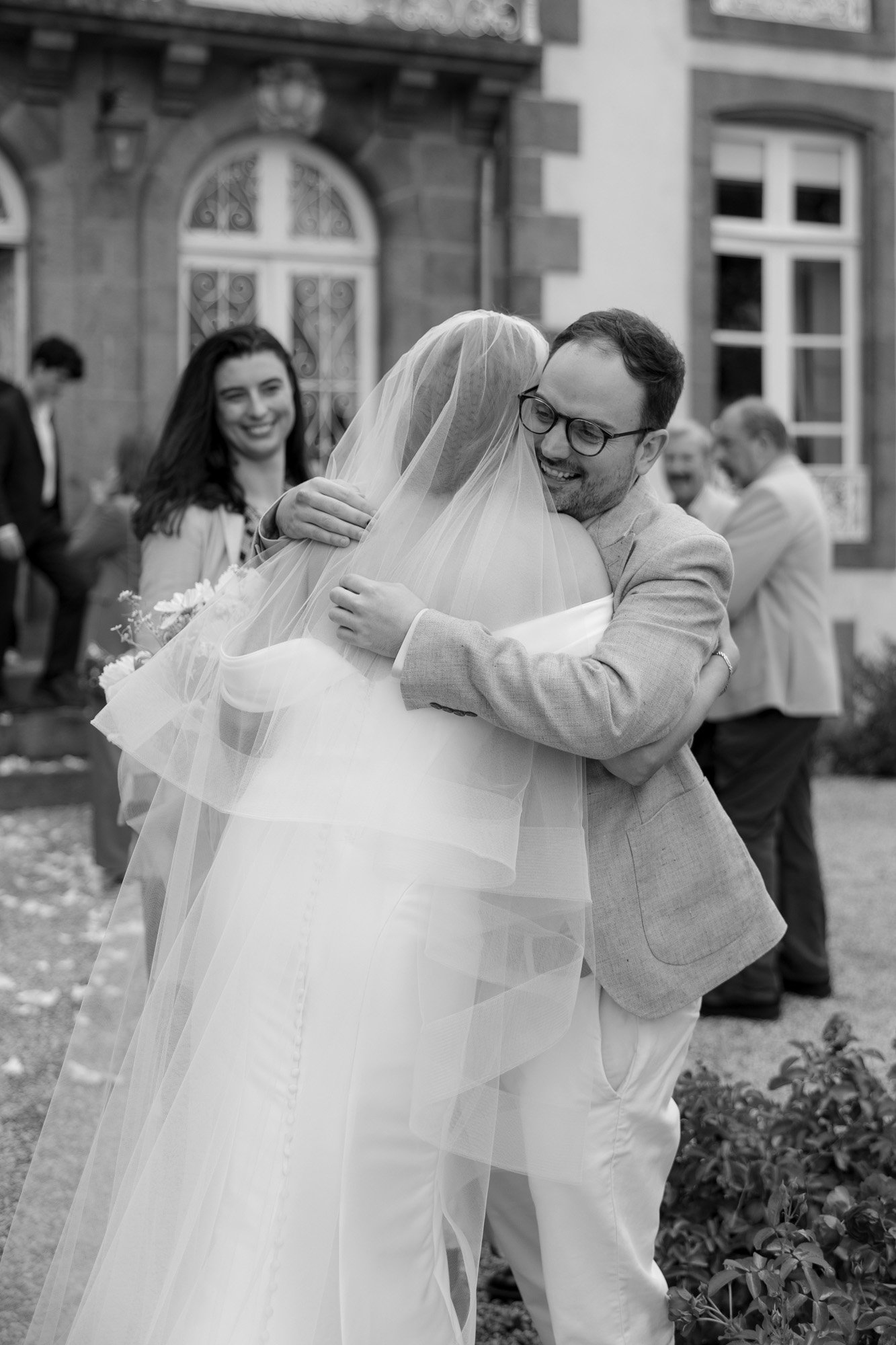 A bride in a wedding dress and veil hugs a smiling man in a light suit outdoors, with guests and a large building in the background.
