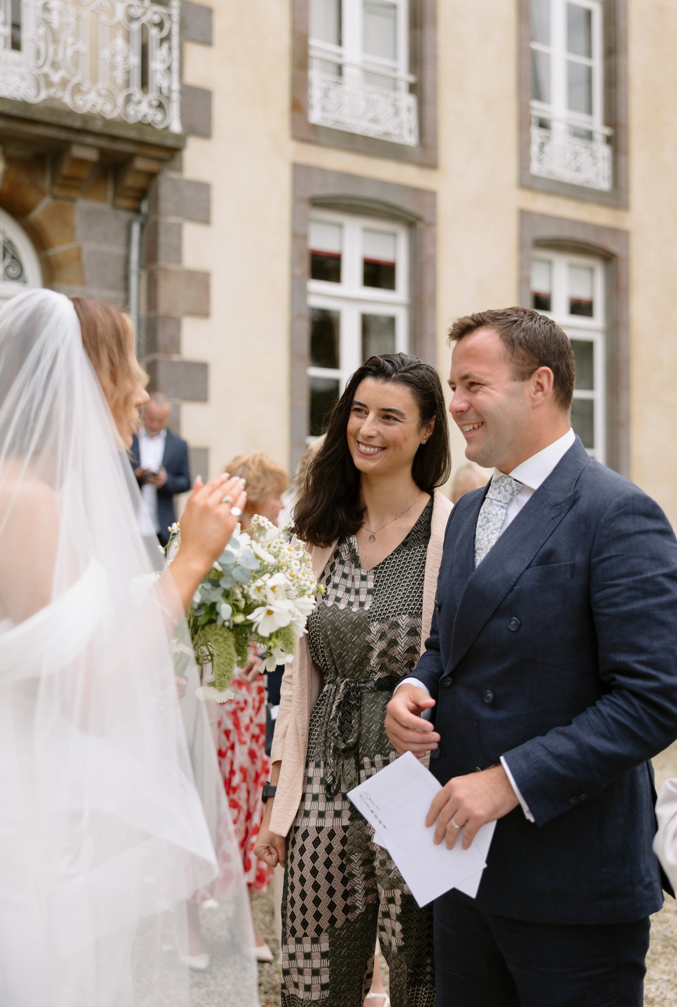 A bride in a veil talks with a woman and a man holding papers outside a building with large windows and decorative railings.