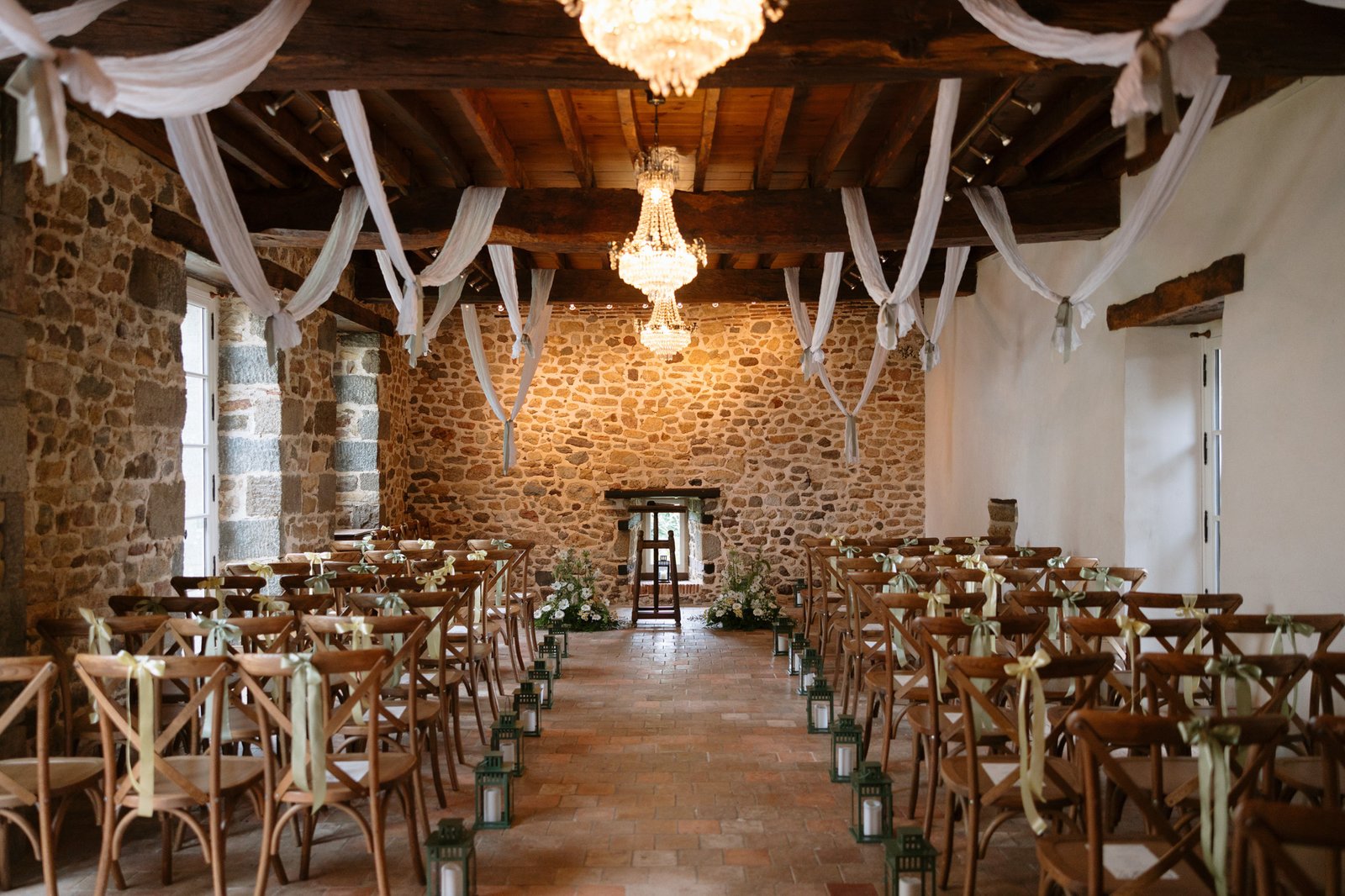 A rustic indoor wedding ceremony setup with wooden chairs, white drapes, chandeliers, floral arrangements, and lanterns lining the aisle in a stone-walled room.