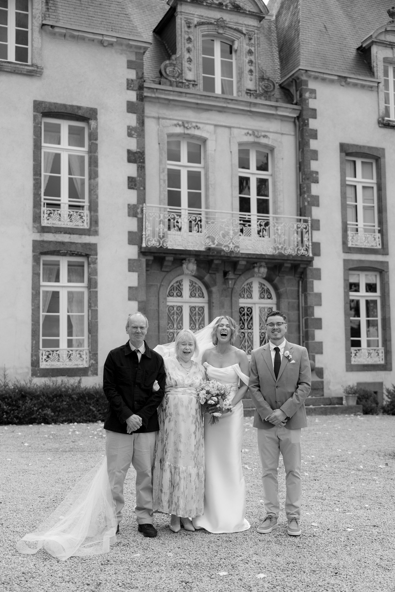 Four people stand posing for a photo in front of a large stone building; one woman wears a wedding dress and veil, others wear formal attire.