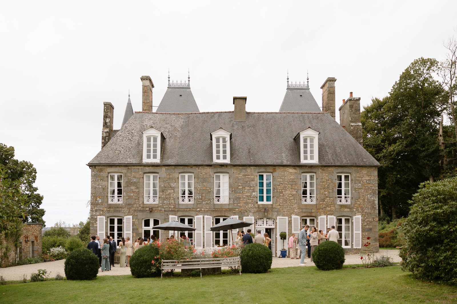A group of people gather in front of a large stone chateau with tall windows and a sloped roof on an overcast day.