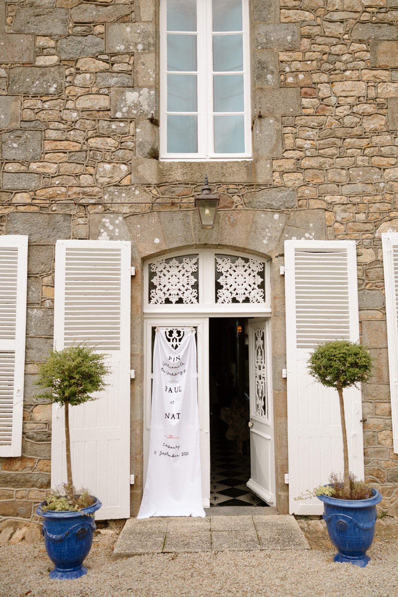 Stone building facade with white shutters and double doors, a decorative wedding banner hanging in the entrance, and two potted topiary trees in blue pots on either side.