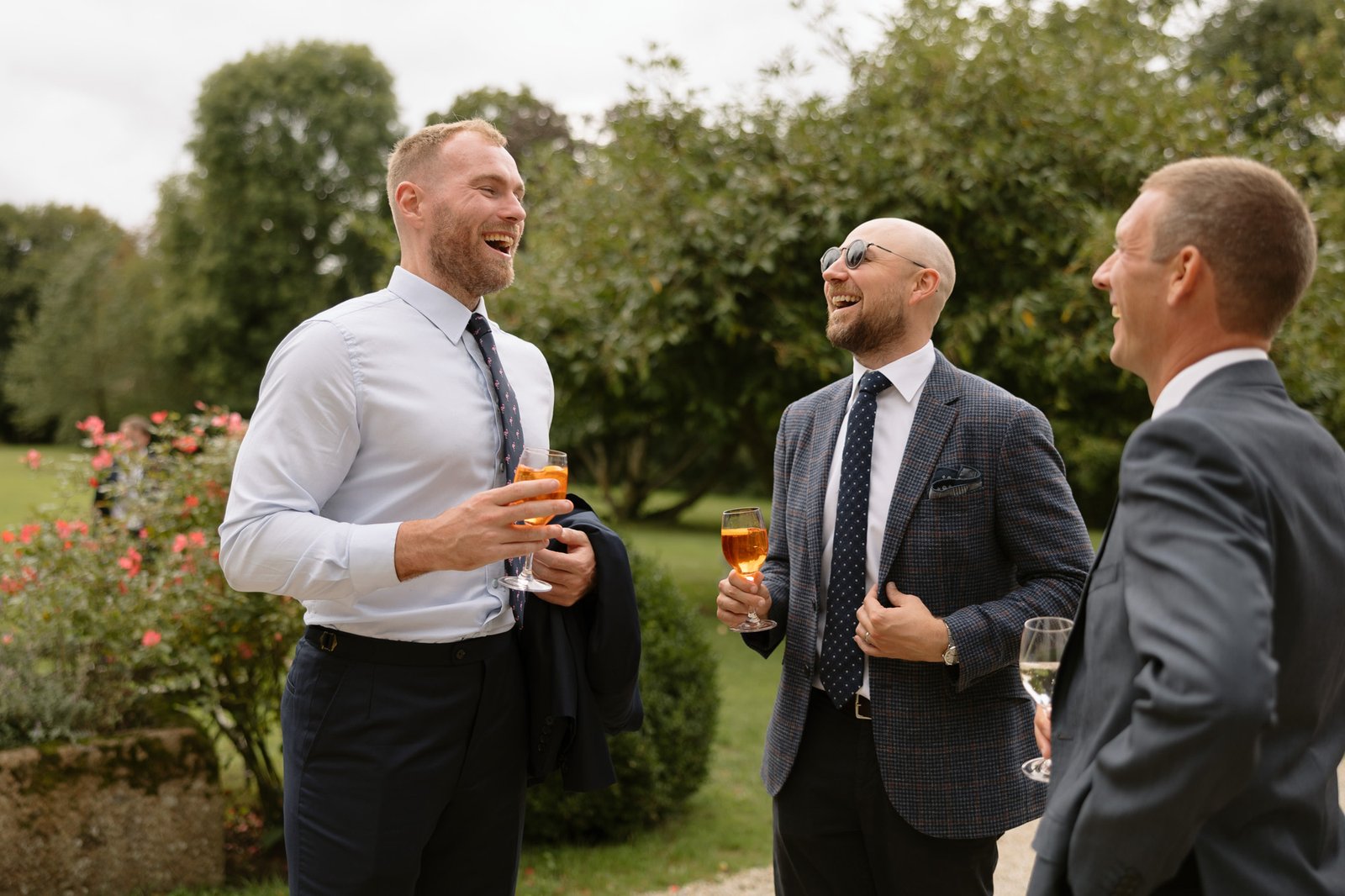 Three men in suits stand outdoors, holding drinks and laughing together, with trees and greenery in the background.