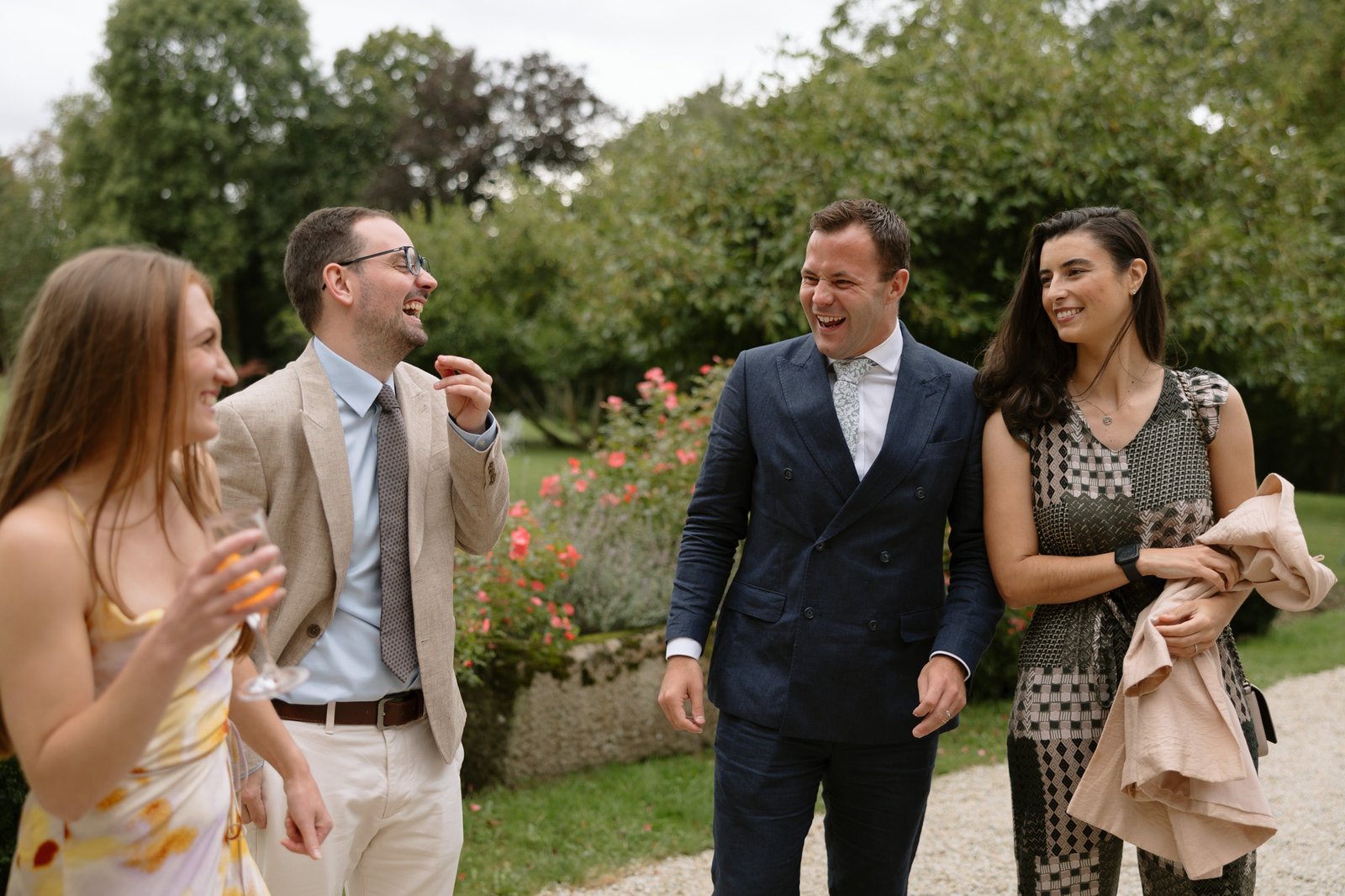 Four adults dressed in semi-formal attire stand outdoors, smiling and talking, with trees and flowers in the background on a cloudy day.
