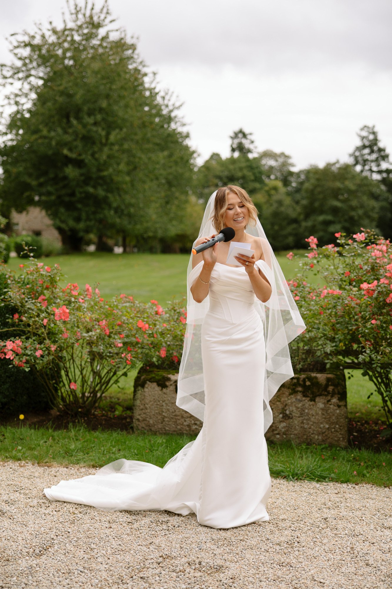 A bride in a white wedding dress and veil stands outdoors holding a microphone and a piece of paper, with pink flowers and greenery in the background.