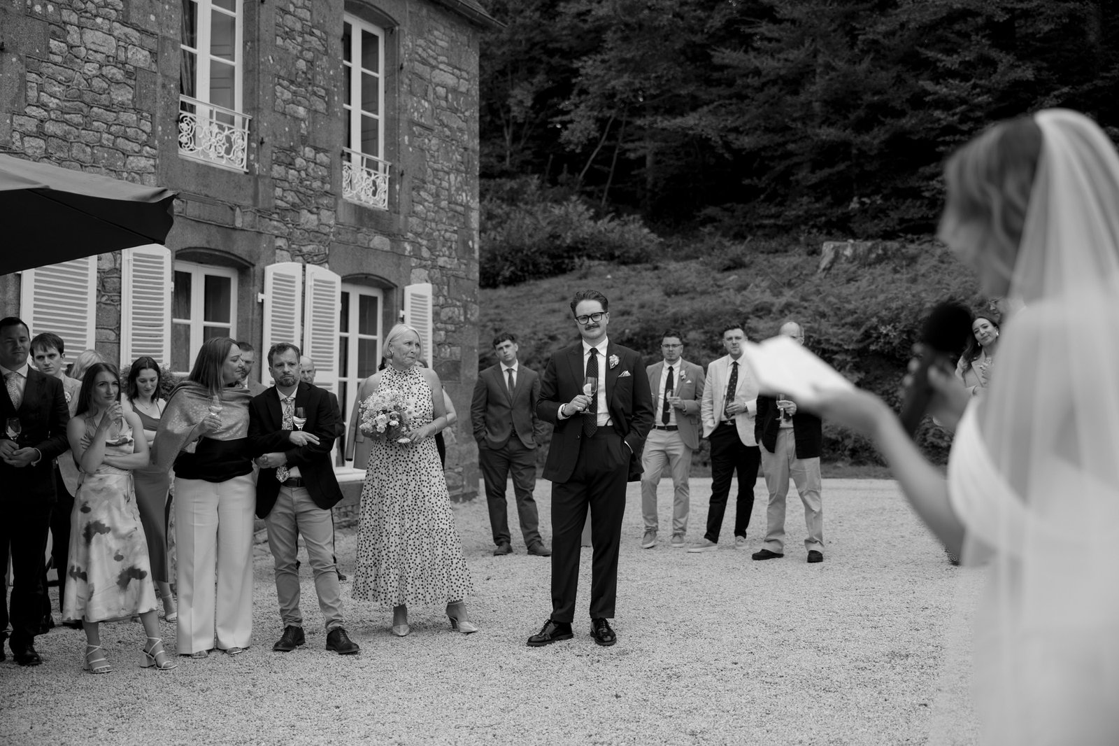 A bride speaks into a microphone while wedding guests stand outside a stone building, listening attentively.