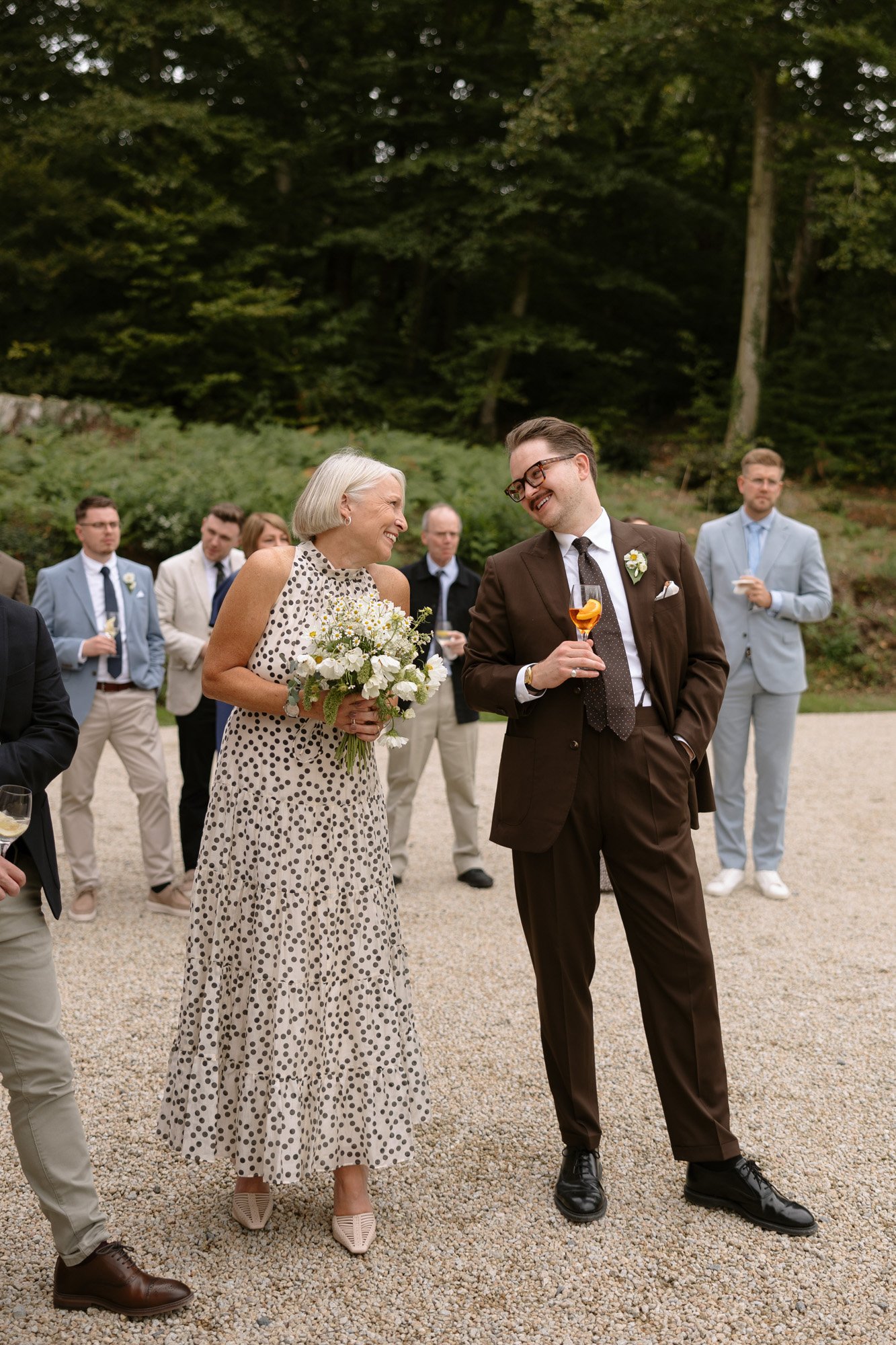 A woman in a polka dot dress holding a bouquet and a man in a brown suit holding a drink are smiling outdoors among a group of formally dressed people.