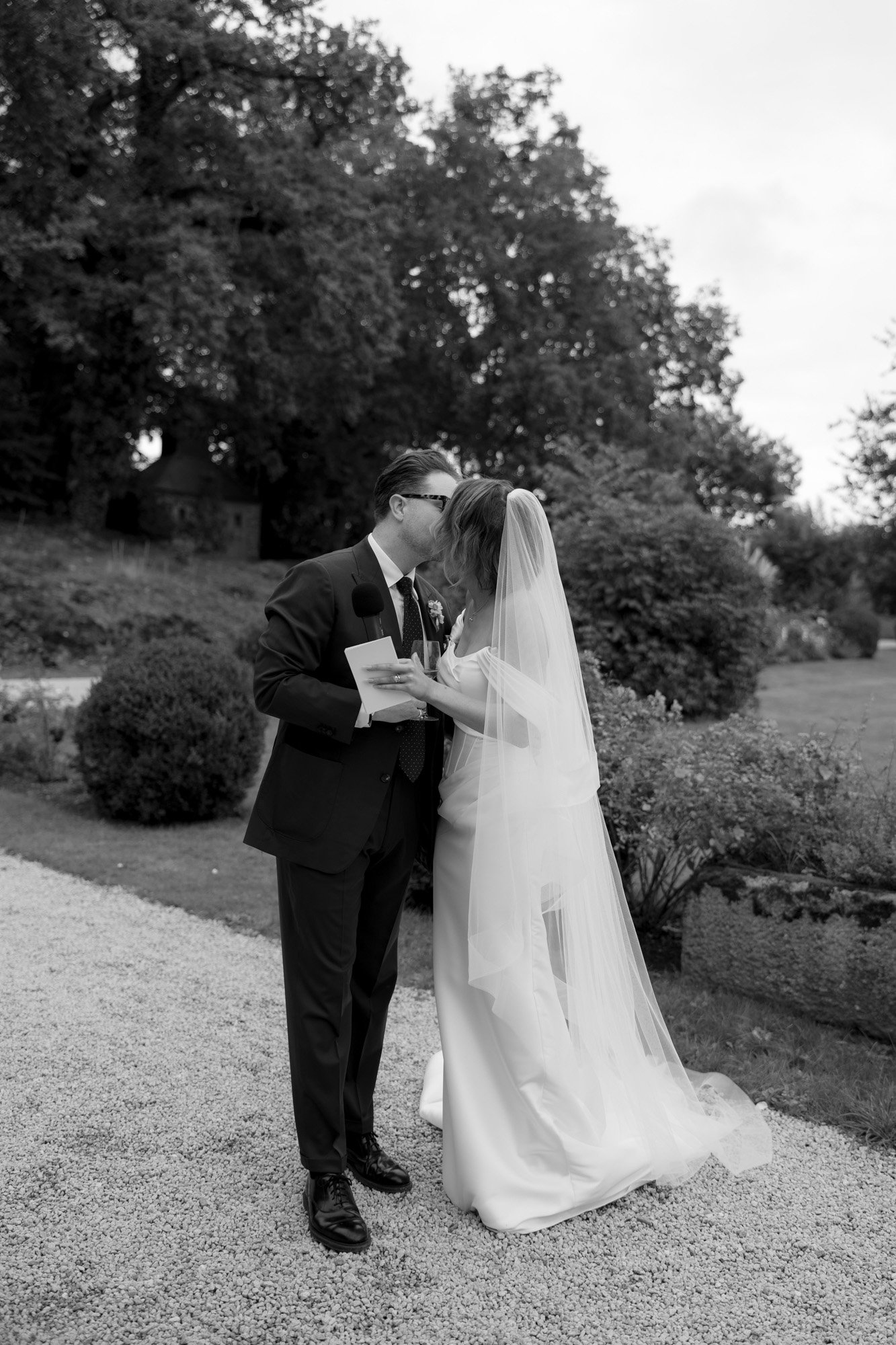 A bride and groom kiss outdoors, with the bride holding a piece of paper and a microphone; trees and shrubbery appear in the background.