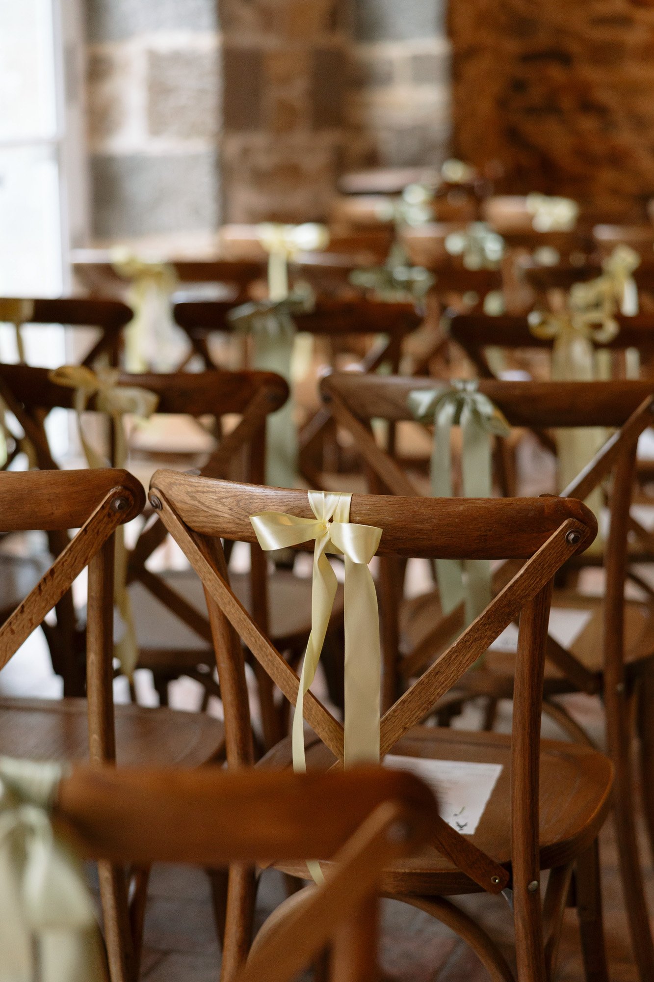 Rows of wooden chairs with light-colored ribbons tied on the backs, arranged indoors near stone walls, likely set up for an event or ceremony.