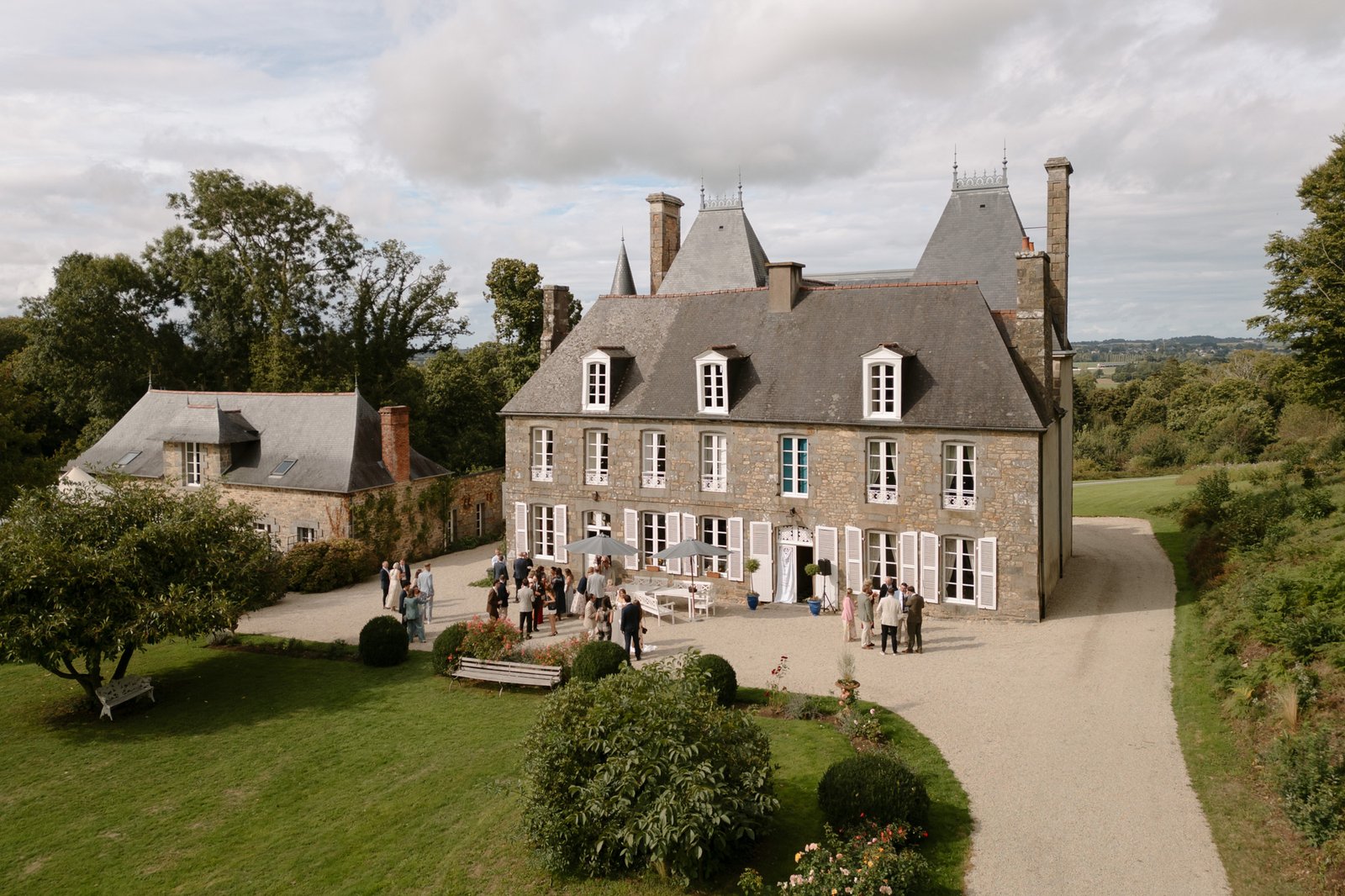 A group of people gathers outside a large stone manor with steep roofs and tall chimneys, surrounded by green gardens and trees under a partly cloudy sky.