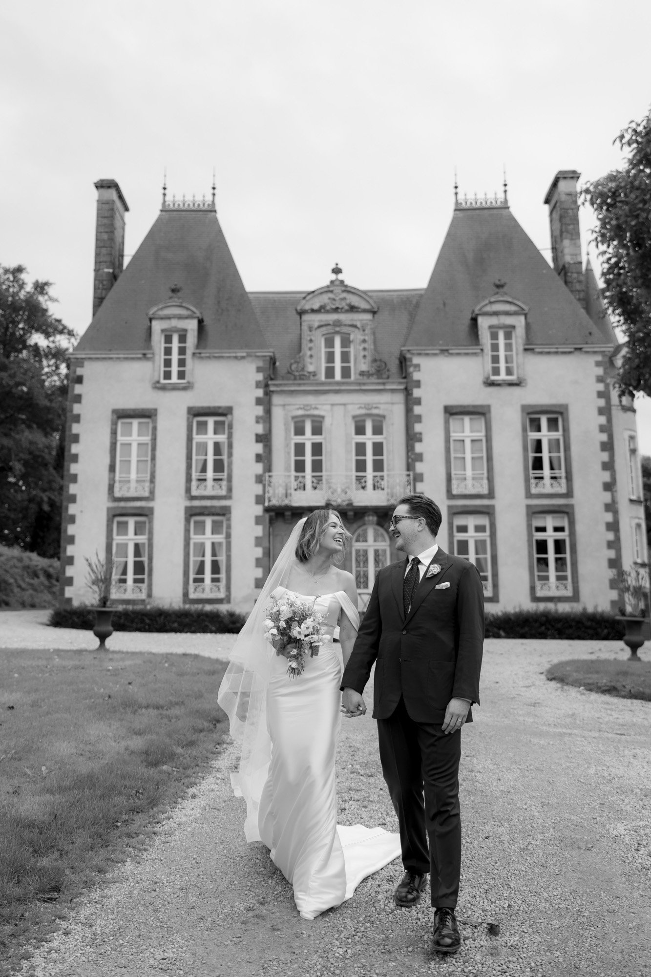 A bride and groom walk hand in hand, smiling at each other, in front of a large historic building with steep roofs.