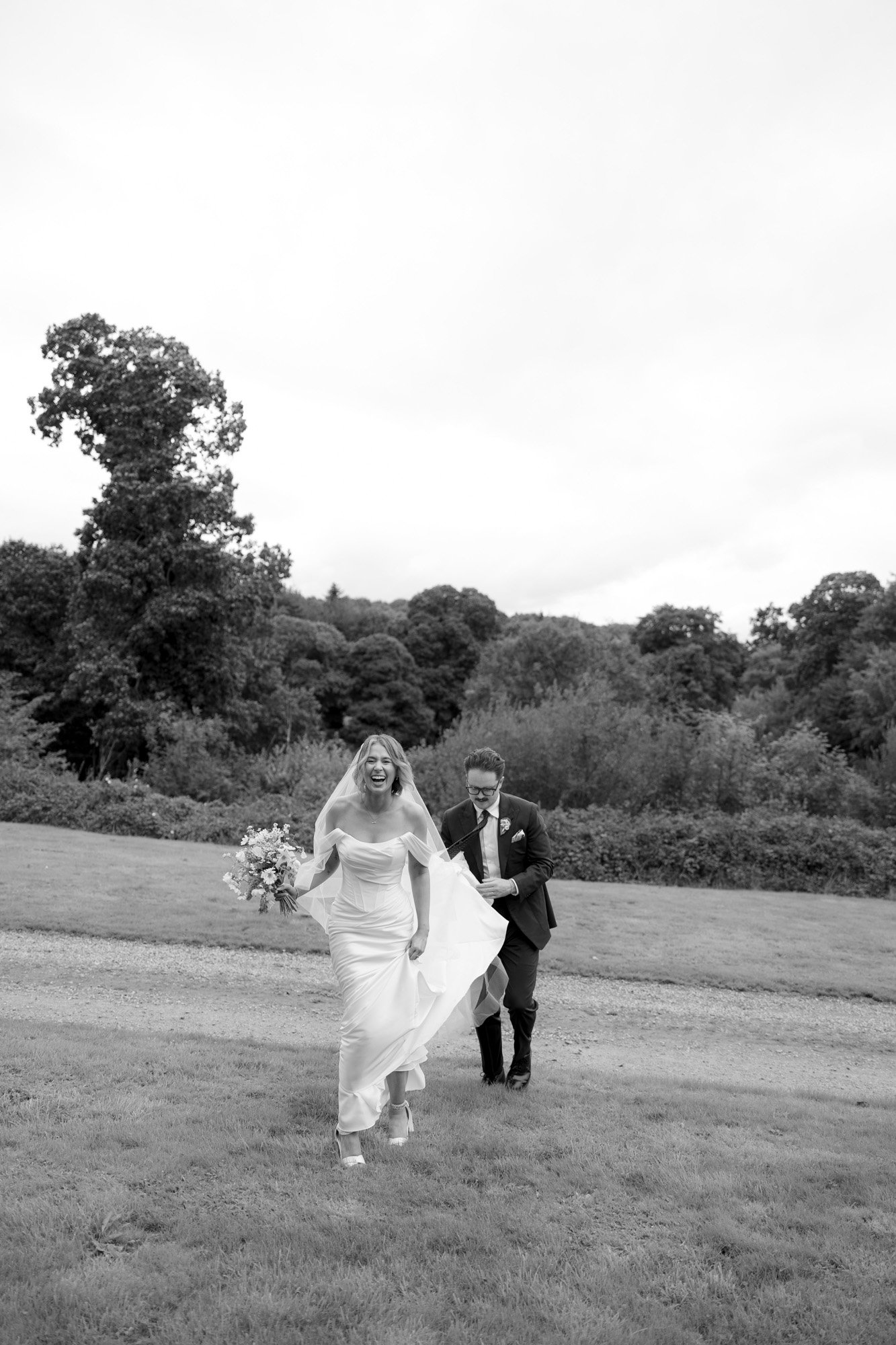 A bride in a white dress holding a bouquet walks on grass, followed by a man in a suit, with trees and shrubs in the background.
