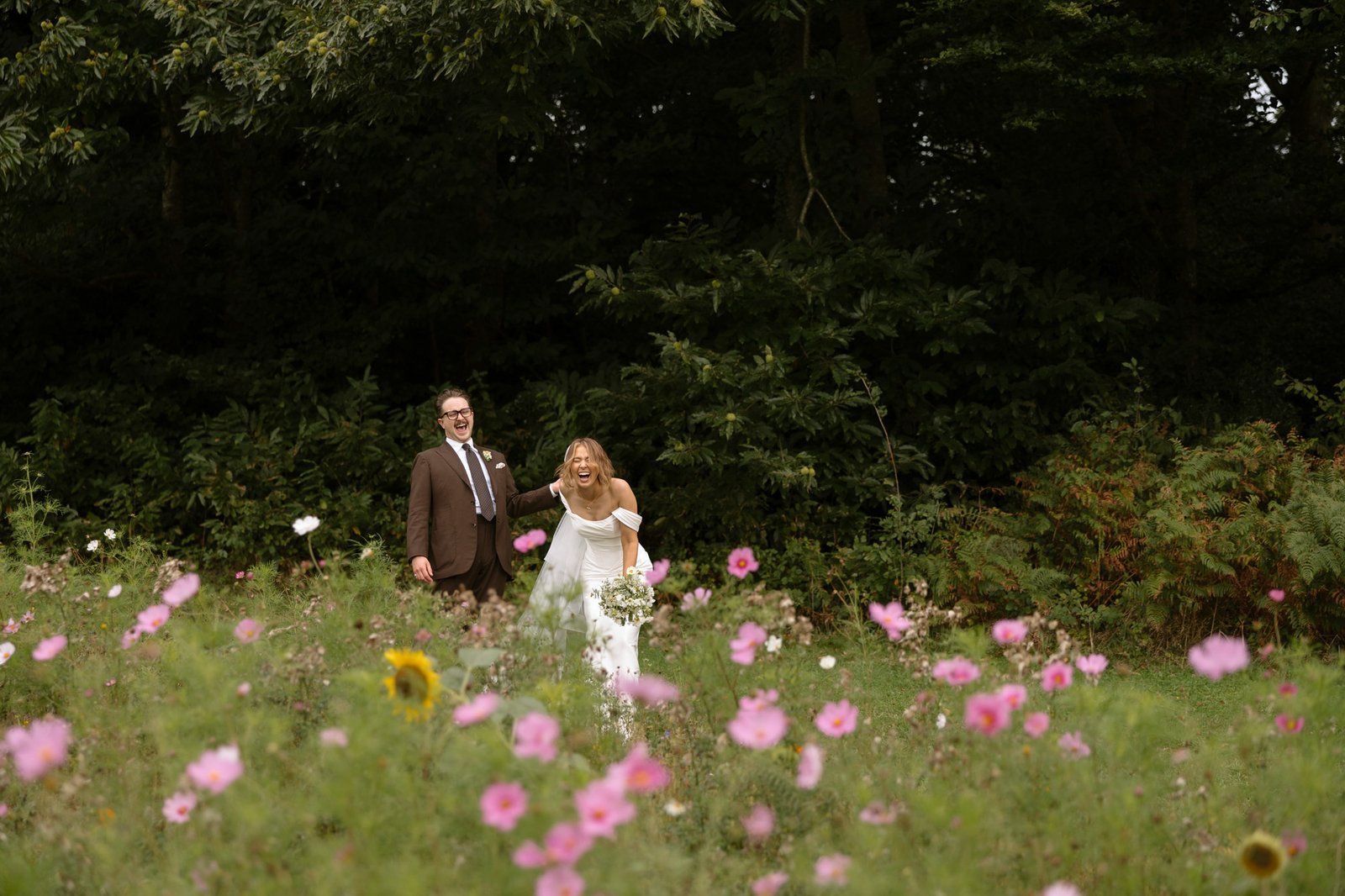 A bride and groom stand smiling in a field of wildflowers with trees in the background.