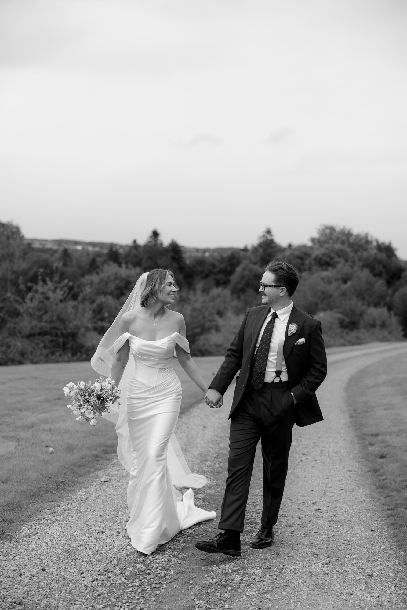 A bride and groom holding hands and smiling at each other while walking along a gravel path outdoors.