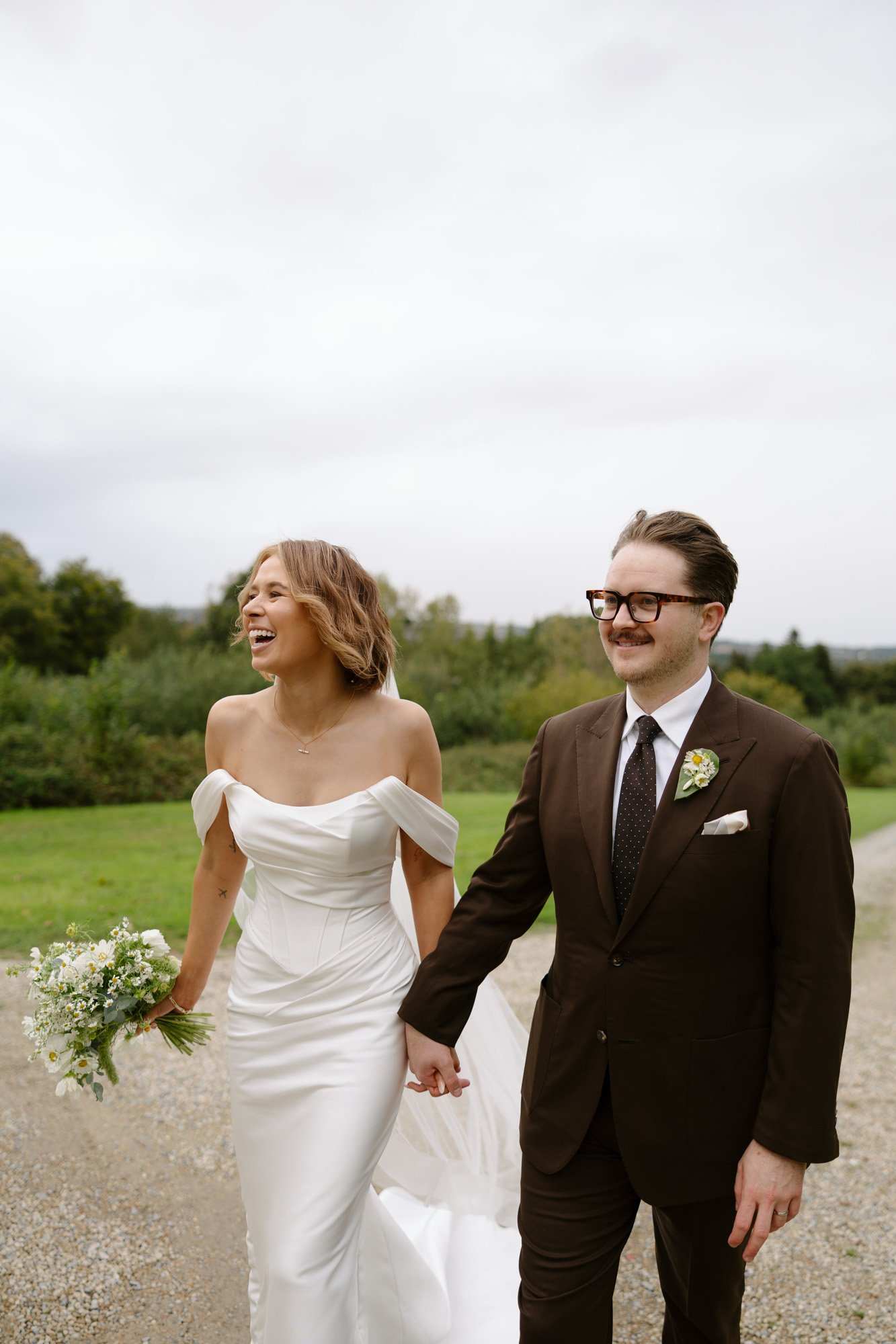 A bride in a white dress and a groom in a brown suit walk hand in hand outdoors, both smiling, with greenery and cloudy sky in the background.