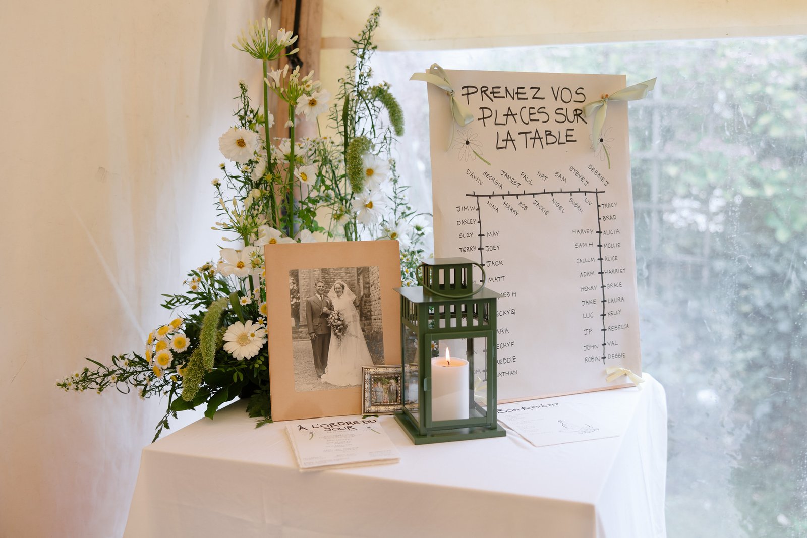 A table displays a floral arrangement, a framed black-and-white wedding photo, a green lantern with a candle, and a seating chart labeled PRENEZ VOS PLACES SUR LA TABLE.