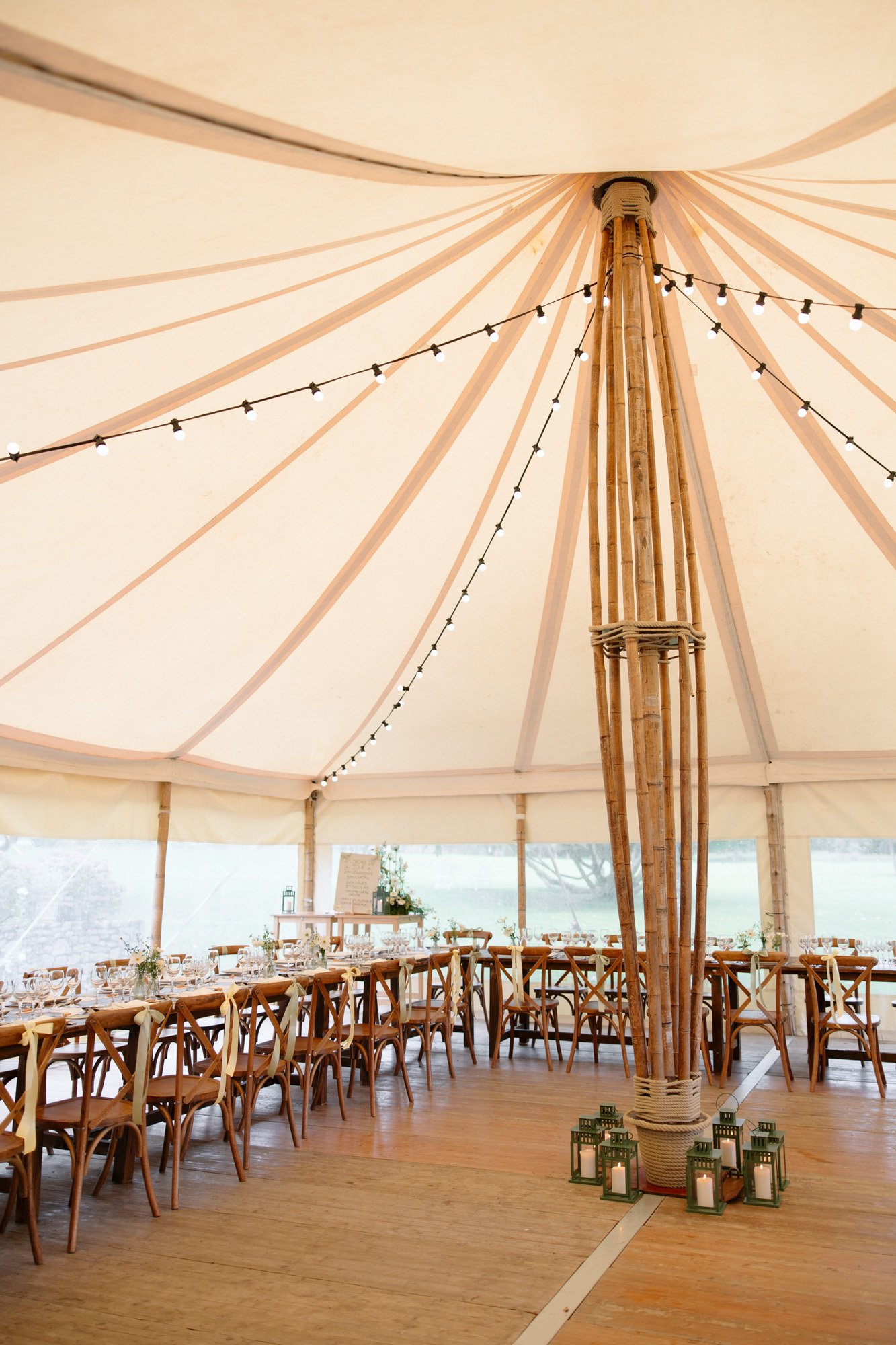 Large tent interior with a central wooden pole, strings of lights, long wooden tables with chairs, and green lanterns on the wooden floor.