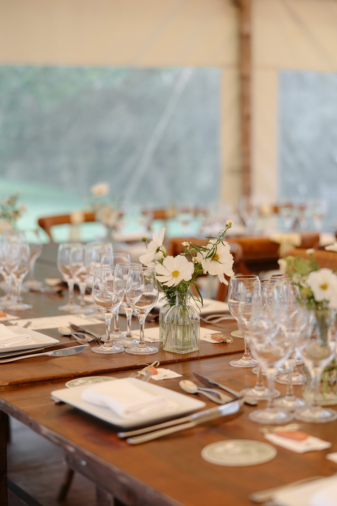 A wooden table set for a formal meal with white plates, cutlery, multiple wine glasses, and small vases of white flowers as centerpieces.