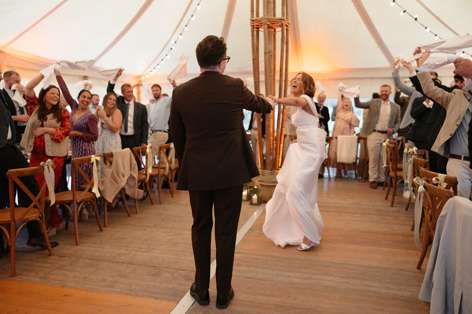 A bride and groom dance down the aisle in a tented venue, as guests stand, smile, and wave napkins in celebration.