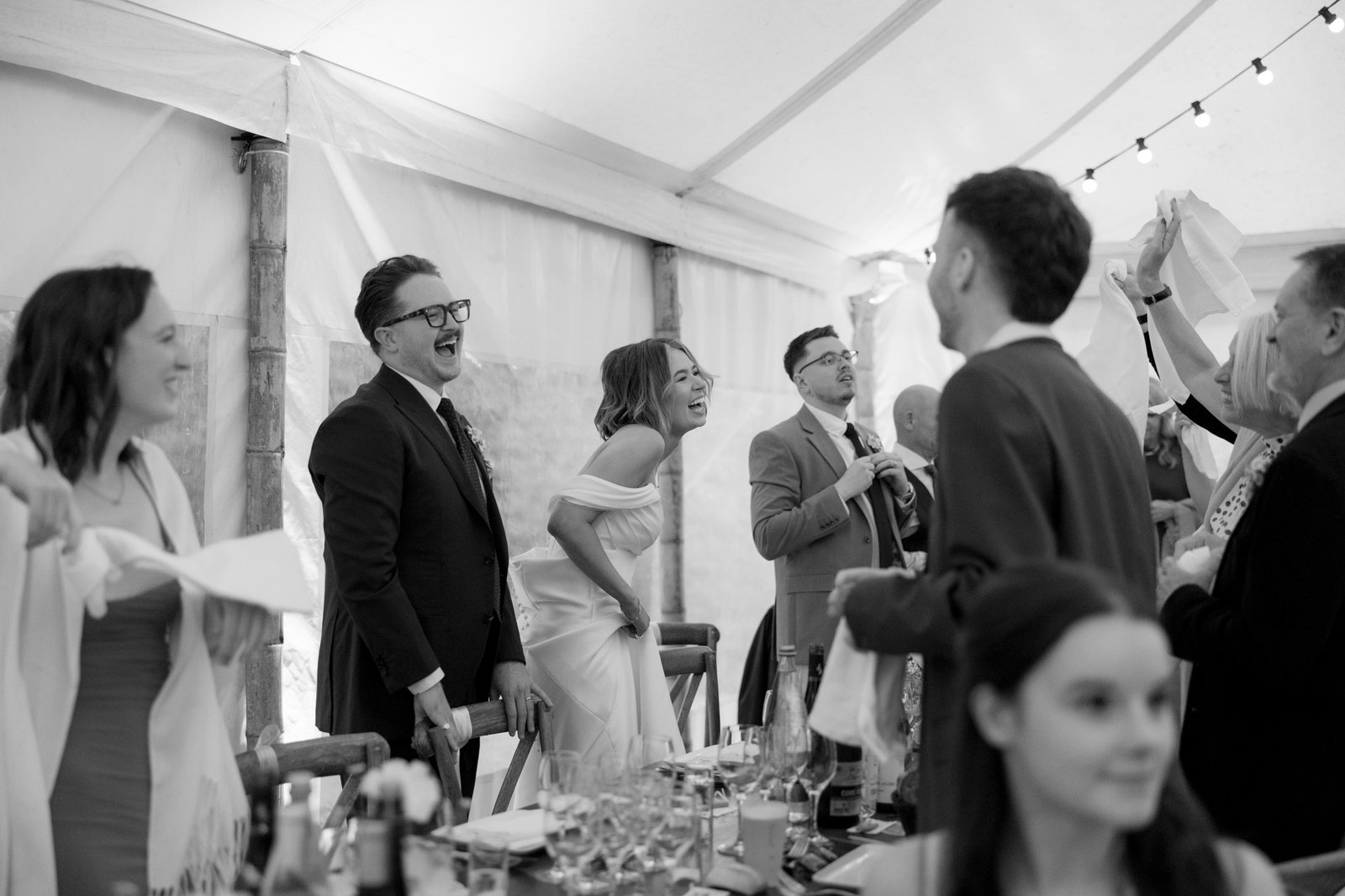 A group of people, including a bride and groom, are standing and laughing inside a tent at a wedding reception. Tables with glasses and bottles are visible in the foreground.