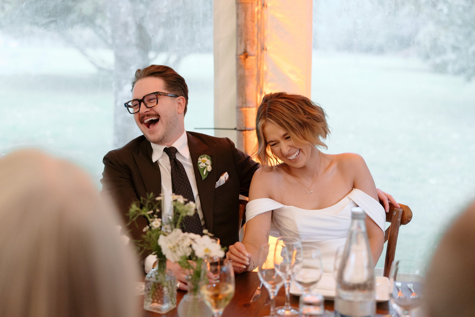 A bride and groom sit together at a table, smiling and laughing, surrounded by flowers, glassware, and wedding decor at a reception.
