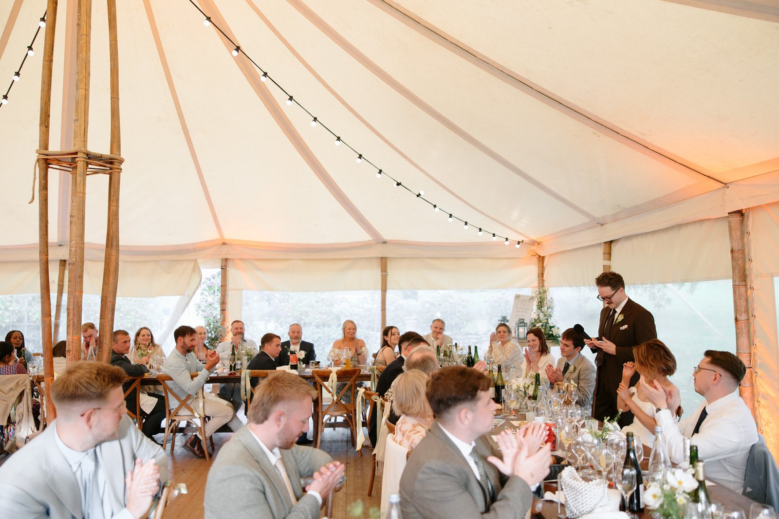 A man stands giving a speech to a seated audience at an indoor event under a large tent, while guests listen and applaud.