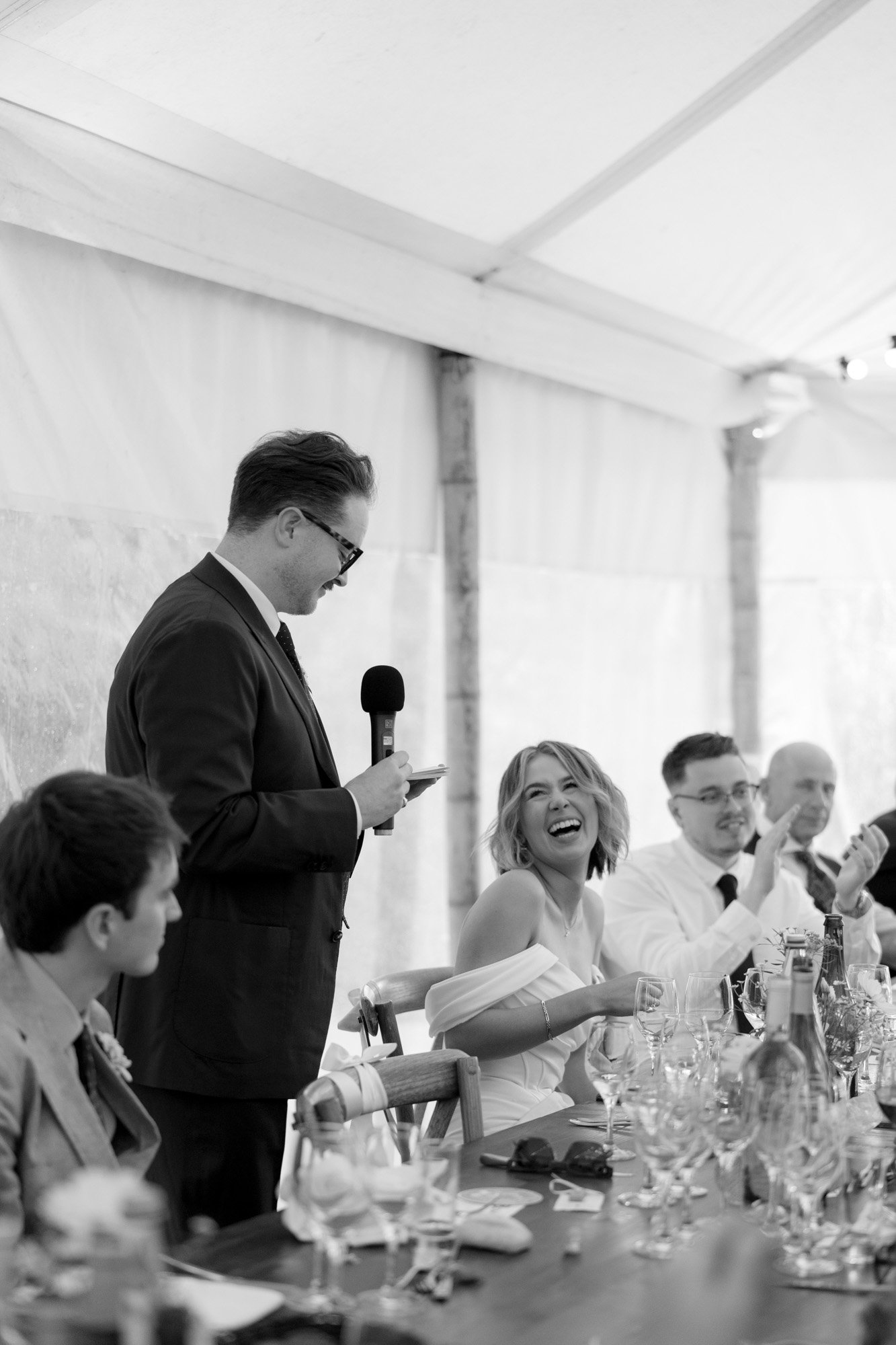 A man in a suit gives a speech with a microphone at a wedding reception while a woman in a white dress smiles and guests sit around the table.